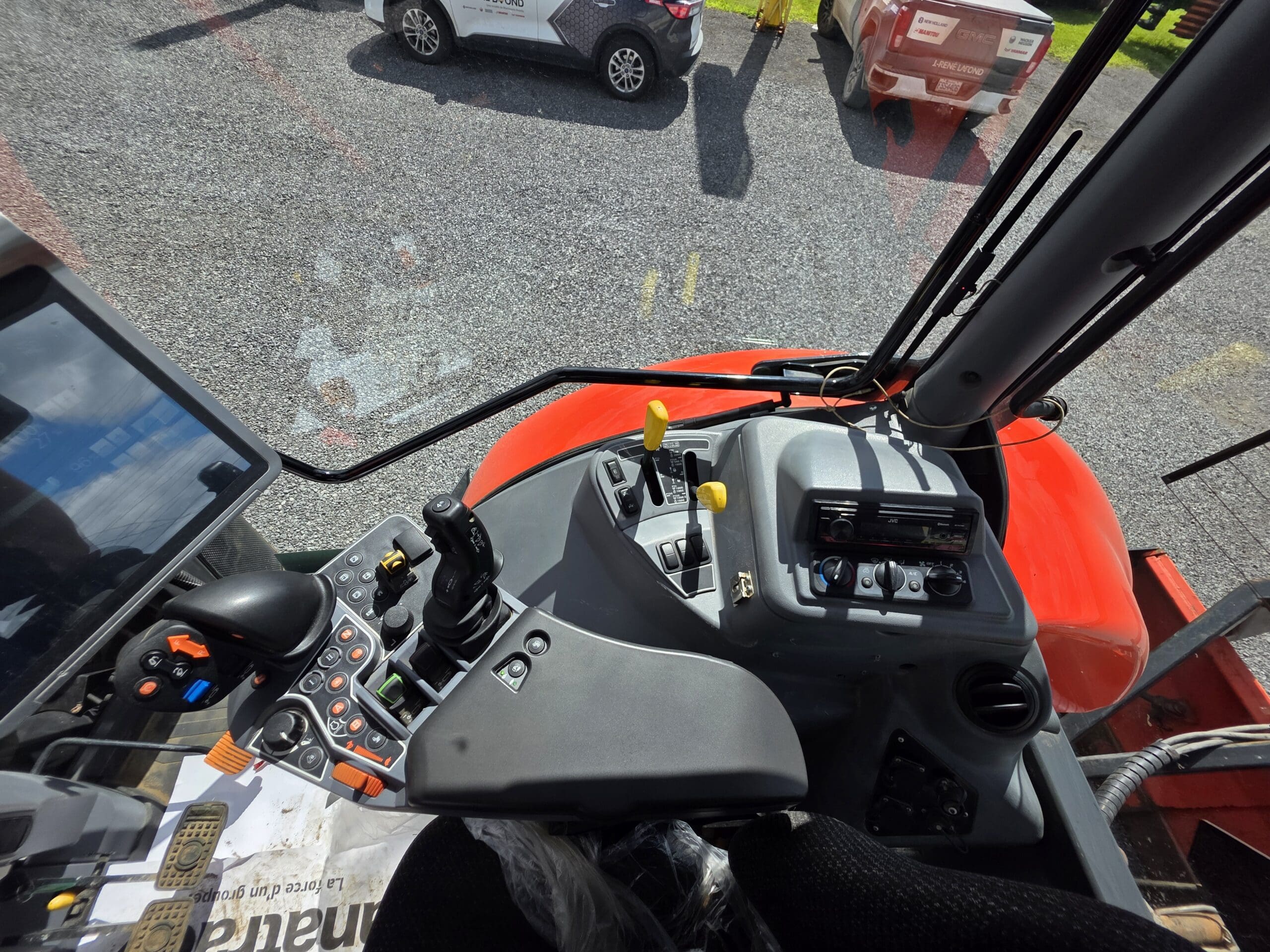Vue de l'intérieur d'une cabine de tracteur montrant le panneau de commande, les leviers, le joystick, les boutons et un écran tactile. Le pare-brise offre une vue sur les véhicules garés sur un terrain en gravier à l'extérieur.