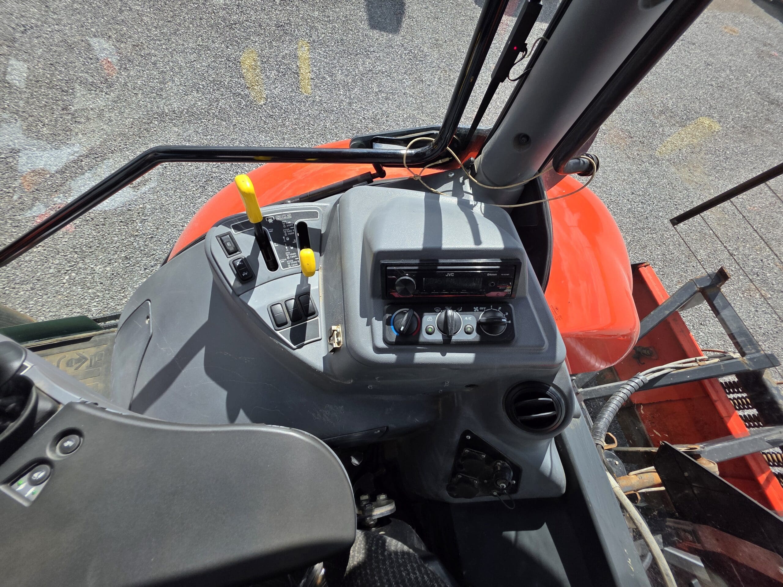 Vue de l'intérieur d'une cabine de tracteur montrant des leviers de commande, des boutons, des bouches d'aération et une radio sur le panneau de commande, avec une partie de l'extérieur rouge et du sol visible à l'extérieur.
