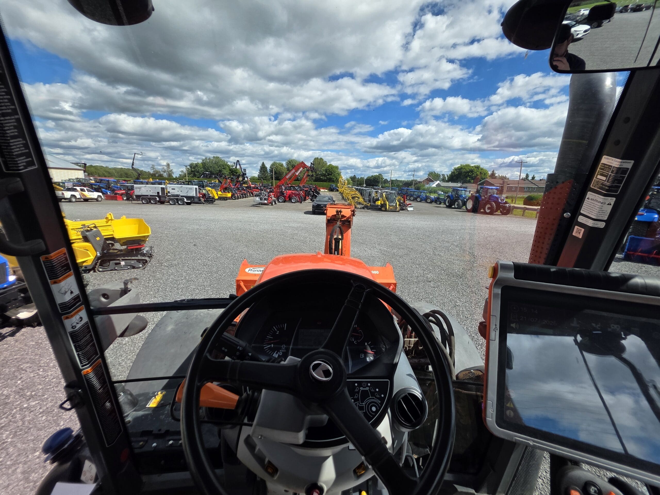 Vue de l'intérieur d'une cabine de tracteur montrant le volant, le tableau de bord et l'écran tactile, donnant sur un terrain rempli de divers équipements de construction et de ferme sous un ciel partiellement nuageux.