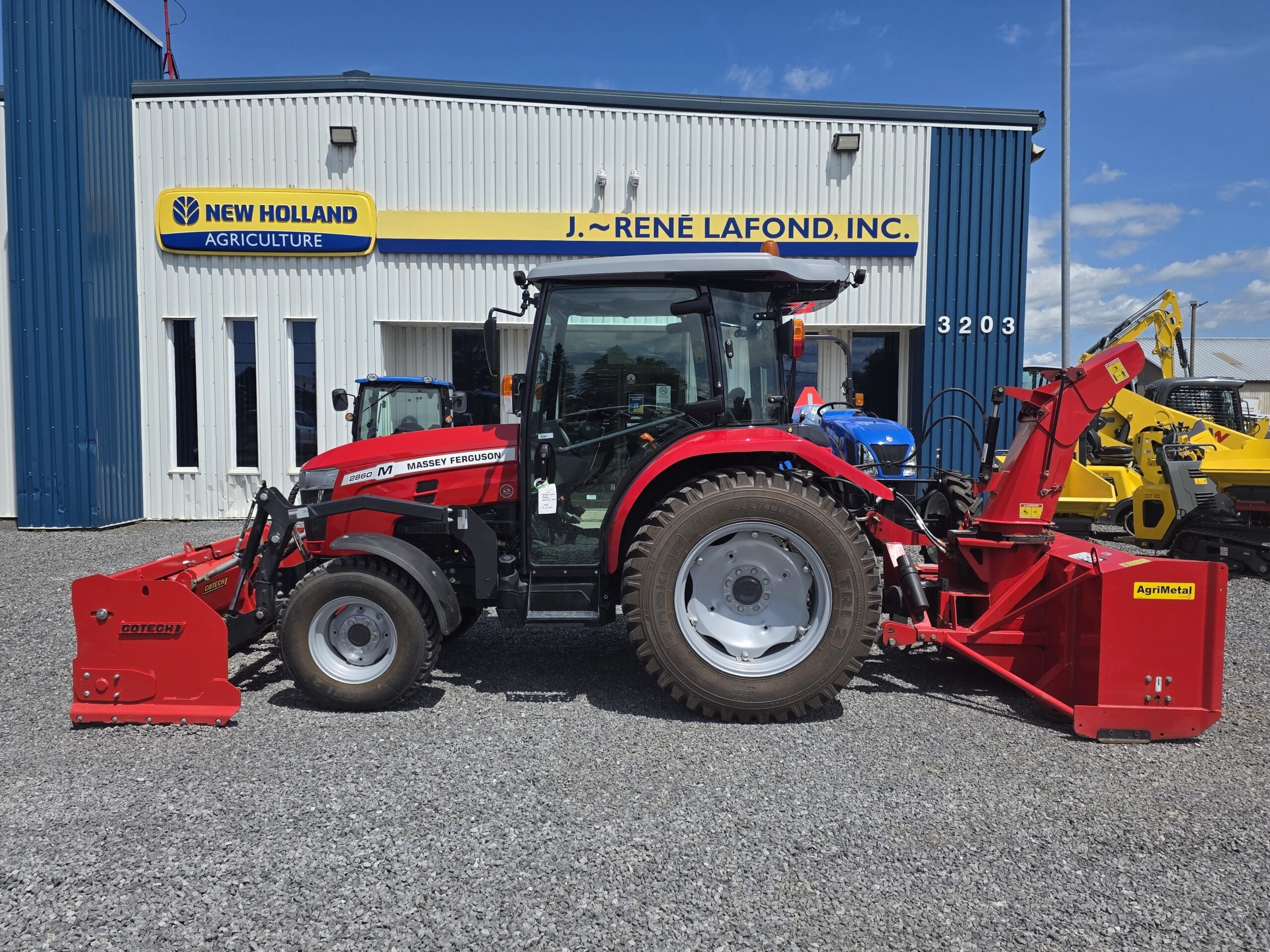 Un tracteur Massey Ferguson rouge avec son matériel agricole rouge est garé devant le bâtiment d'un concessionnaire agricole New Holland sur un terrain en gravier, sous un ciel bleu.