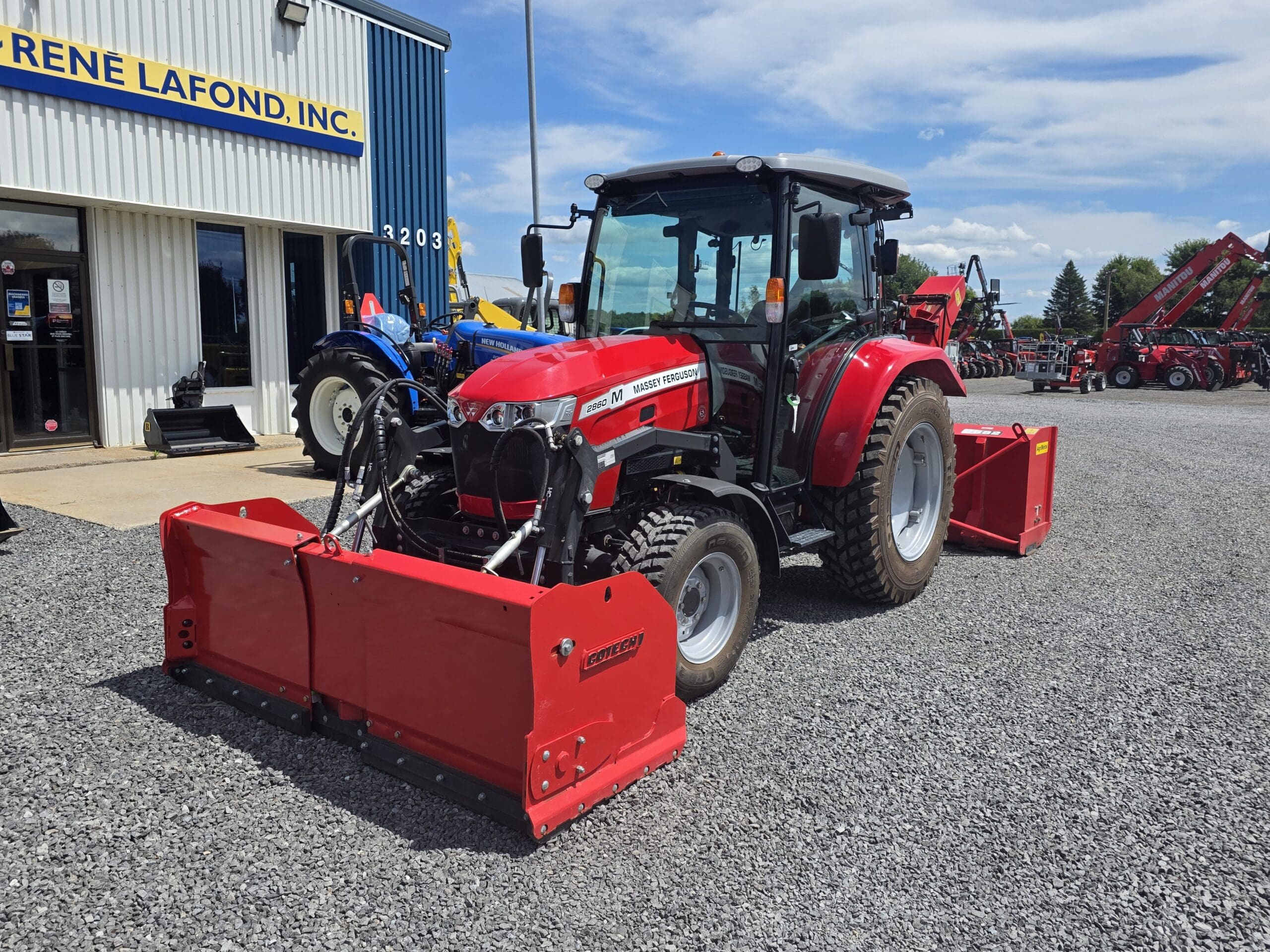 Un tracteur Mahindra rouge équipé de gros outils à l'avant et à l'arrière est garé sur du gravier à l'extérieur d'un bâtiment portant un panneau bleu indiquant 