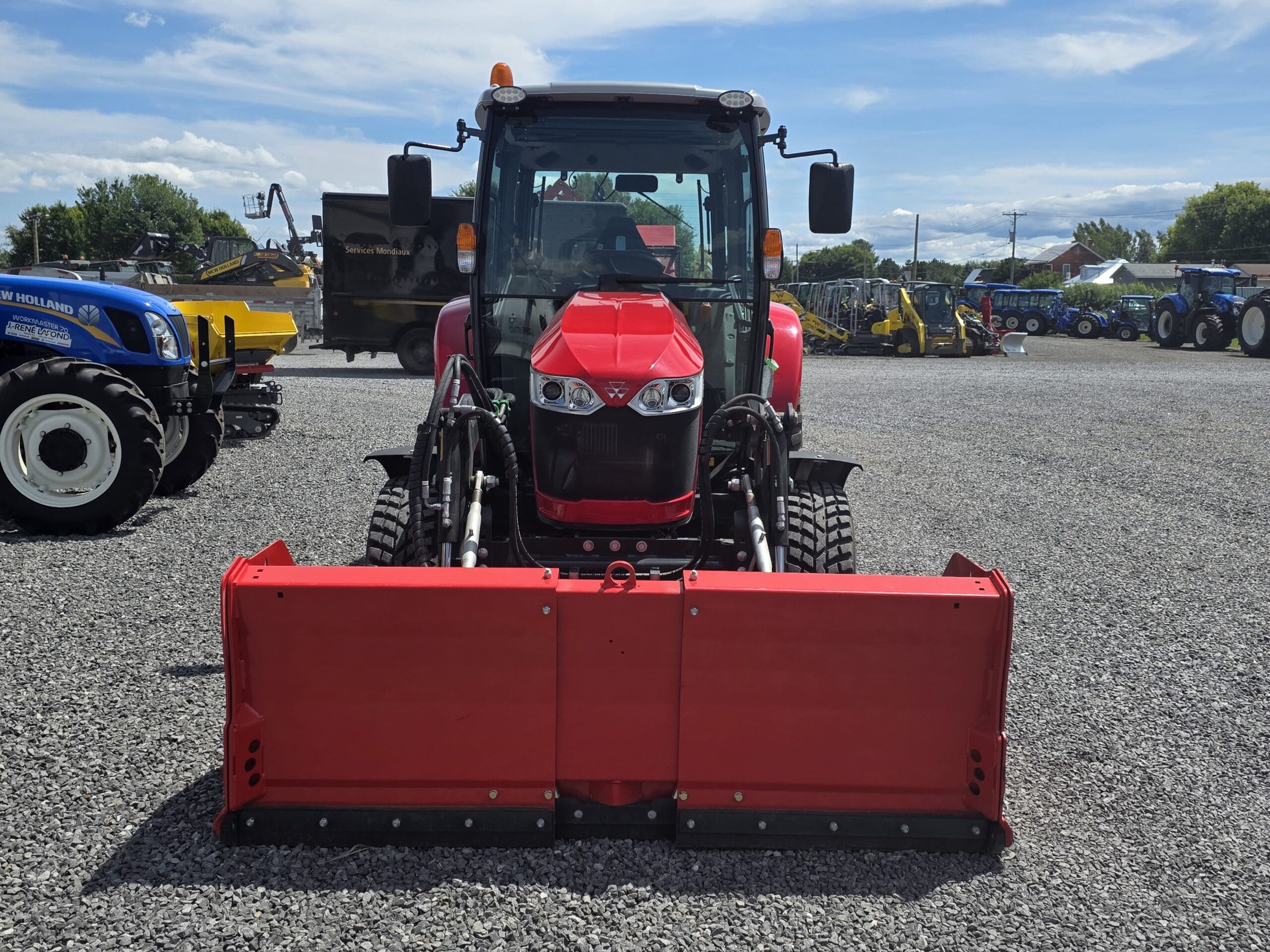 Un tracteur rouge équipé d'un outil frontal est garé sur un terrain en gravier, entouré de divers véhicules de construction et d'exploitation agricole, sous un ciel partiellement nuageux.