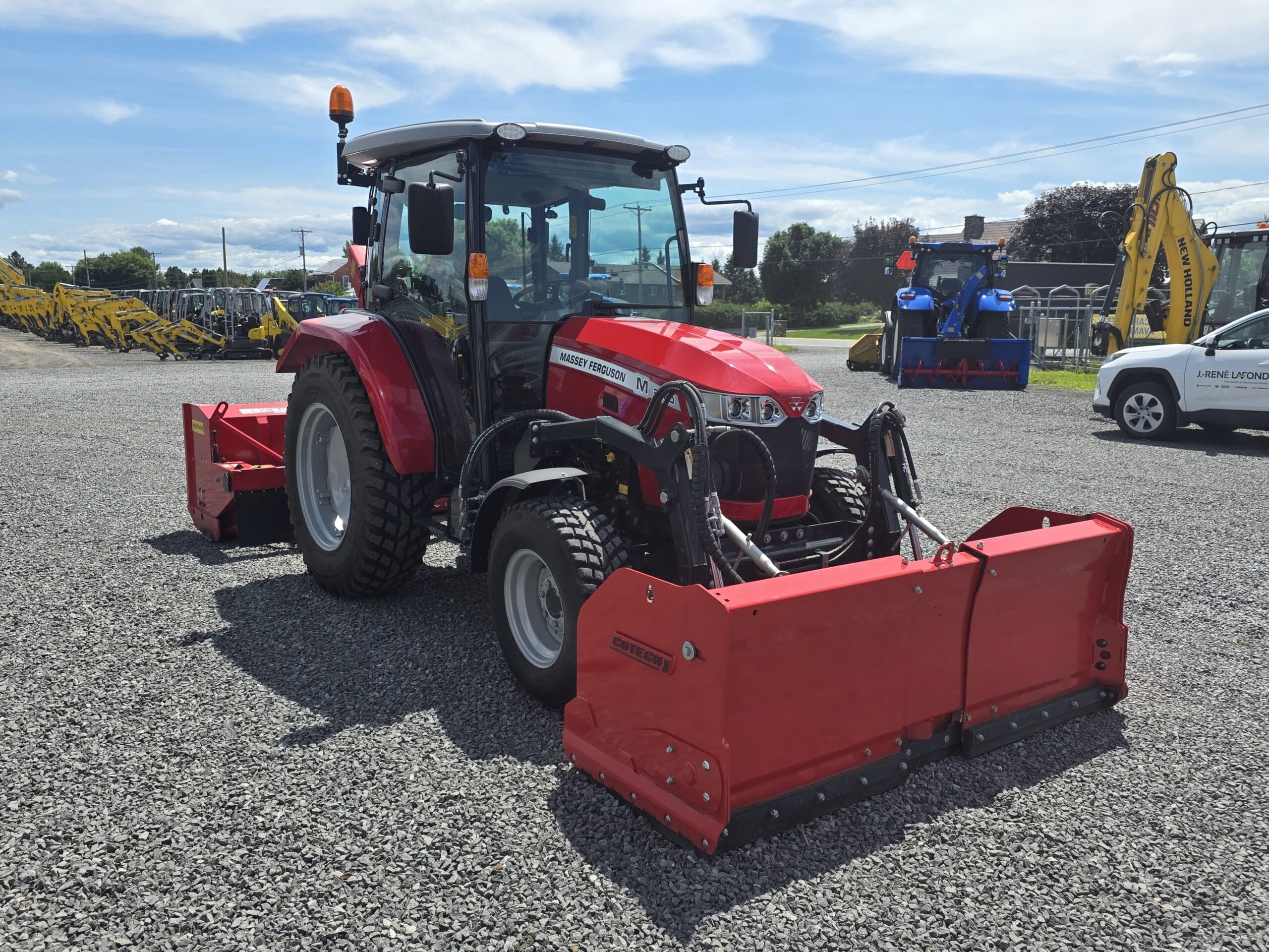Un tracteur compact rouge avec une cabine fermée et de grands outils avant et arrière est garé sur un terrain en gravier sous un ciel partiellement nuageux, avec d'autres machines visibles à l'arrière-plan.