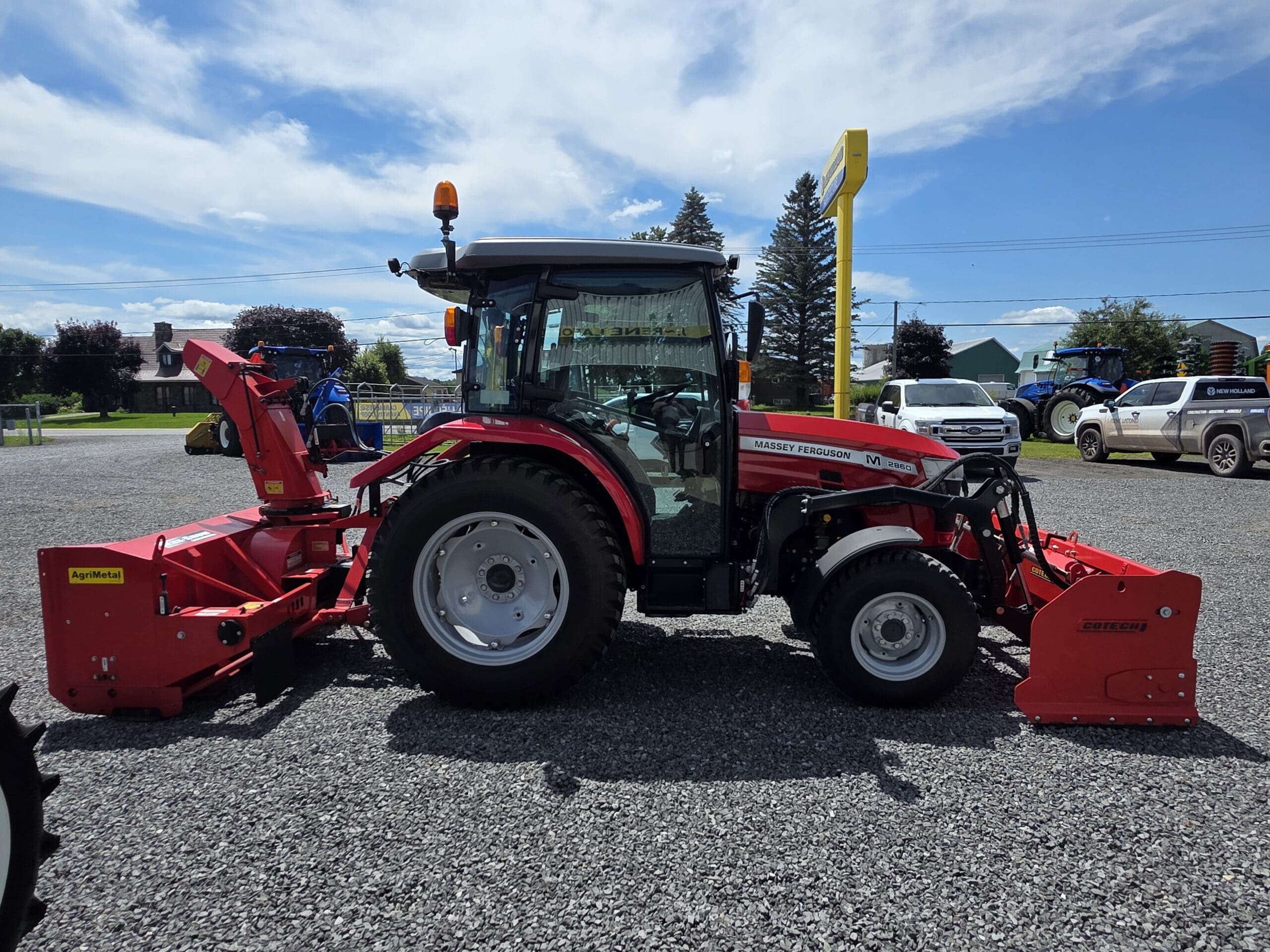 Un tracteur Massey Ferguson rouge avec des machines rouges attachées est garé sur du gravier sous un ciel bleu. Des véhicules et des bâtiments se trouvent à l'arrière-plan.