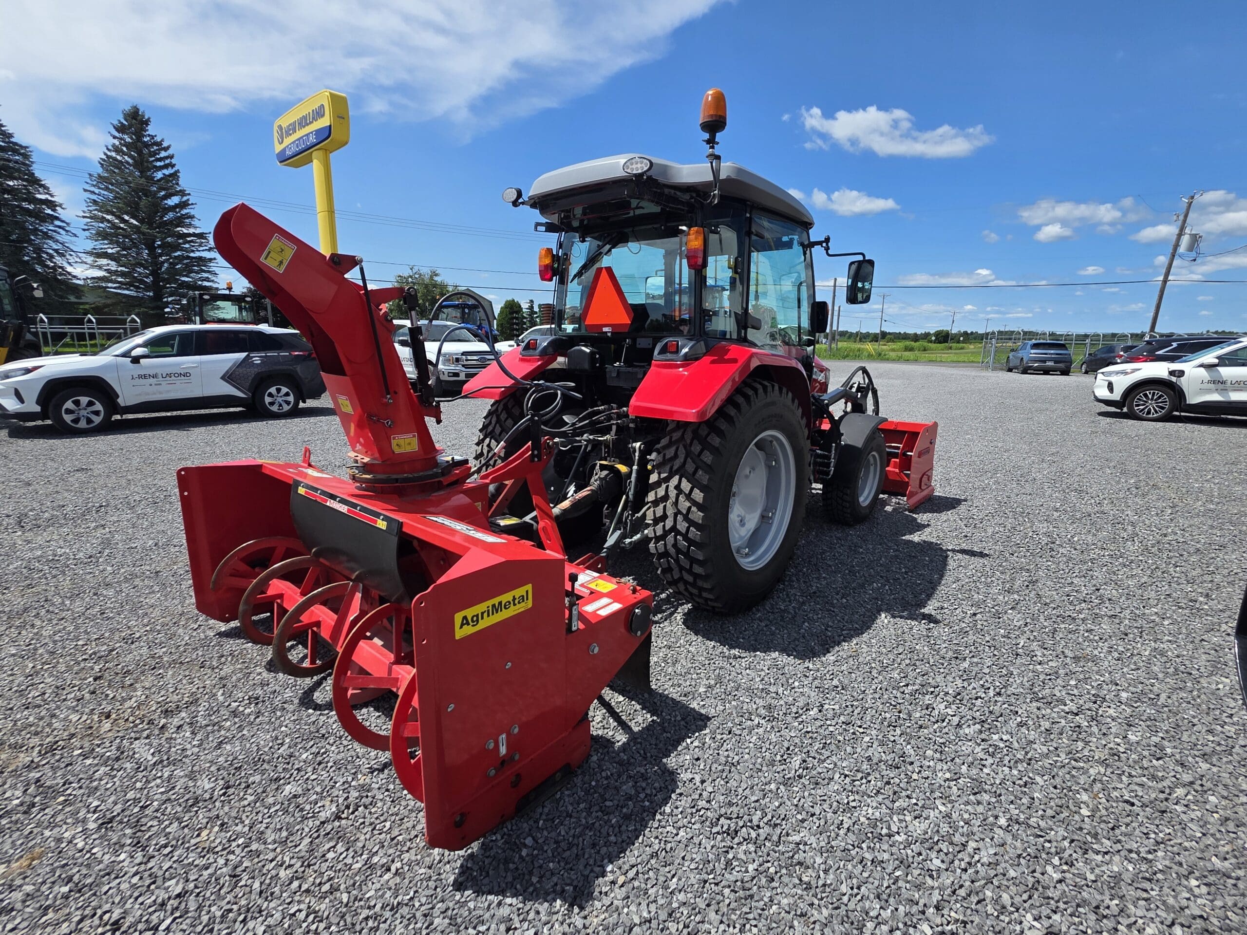 Un tracteur rouge équipé d'une souffleuse à neige rouge est garé sur un terrain en gravier sous un ciel bleu avec des nuages épars. Il y a des voitures et des arbres à l'arrière-plan.
