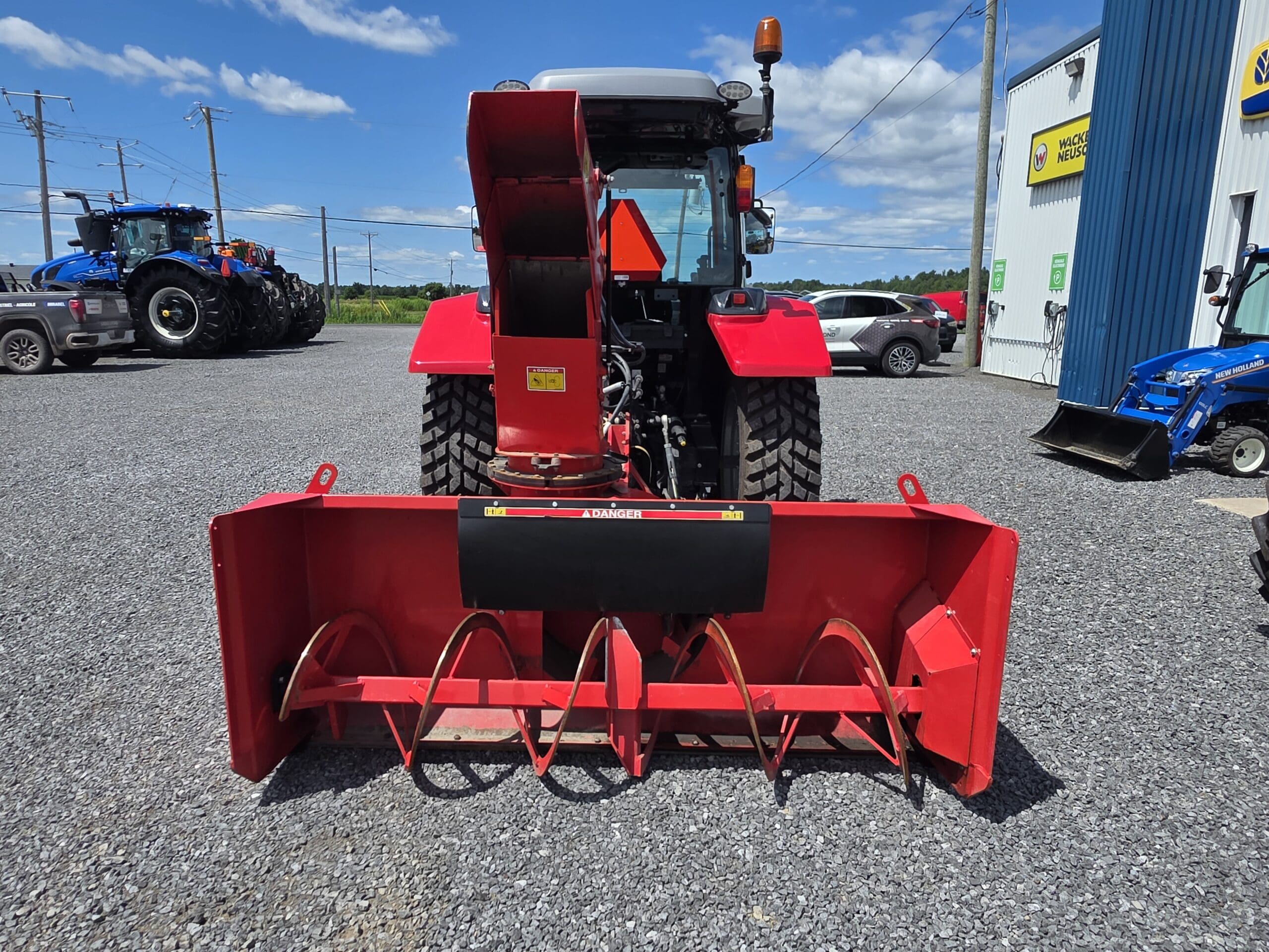 Une souffleuse à neige rouge est montée à l'arrière d'un tracteur, garé sur du gravier à l'extérieur d'un bâtiment au revêtement bleu. D'autres tracteurs et équipements agricoles sont visibles à l'arrière-plan sous un ciel bleu.