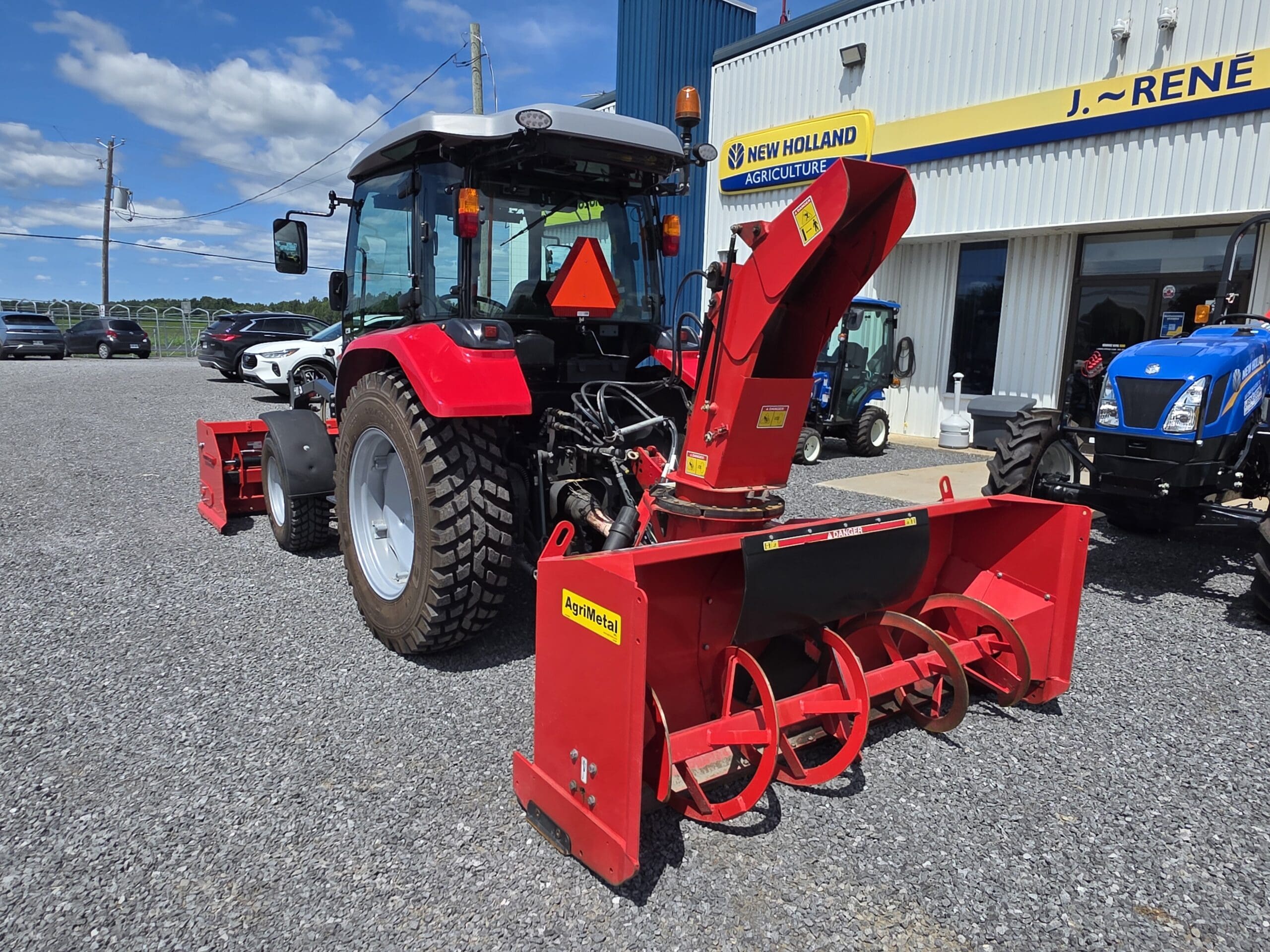 Un tracteur rouge équipé d'une grosse souffleuse à neige rouge est garé devant un concessionnaire New Holland Agriculture par une journée ensoleillée. D'autres tracteurs et véhicules sont visibles à l'arrière-plan.
