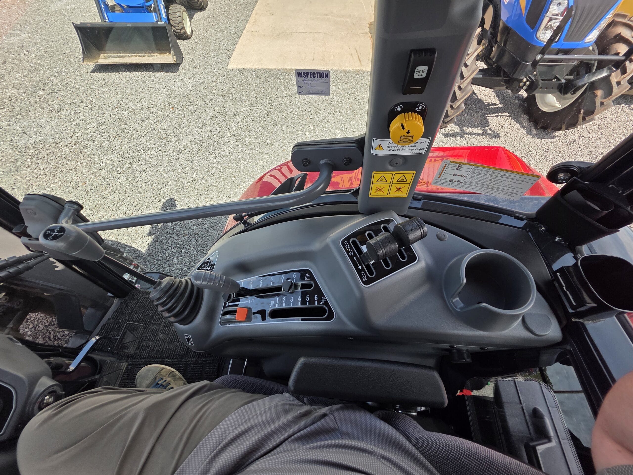 Vue de l'intérieur d'une cabine de tracteur montrant des leviers de commande, des boutons, des étiquettes d'avertissement, un porte-gobelet et une partie de la jambe d'une personne. L'extérieur montre un autre tracteur bleu et un sol en gravier.