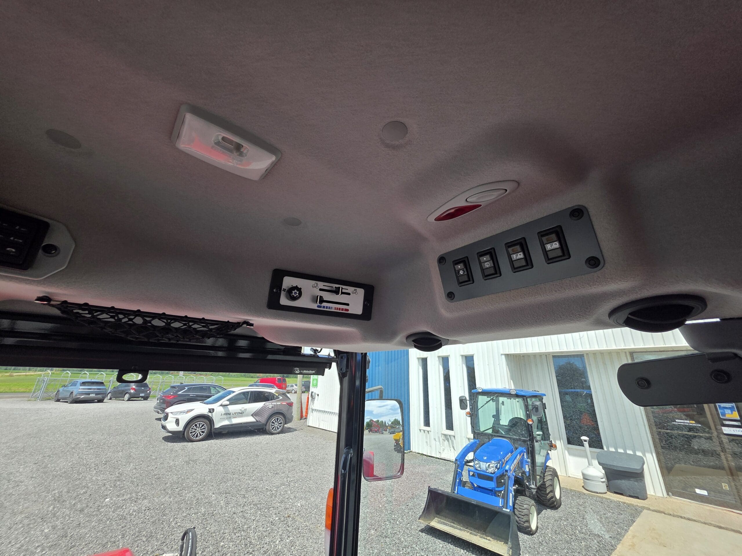 Vue de l'intérieur d'un véhicule montrant ses commandes et interrupteurs au plafond, regardant à travers le pare-brise un tracteur bleu avec un chargeur frontal, garé près d'un bâtiment blanc et de plusieurs voitures.