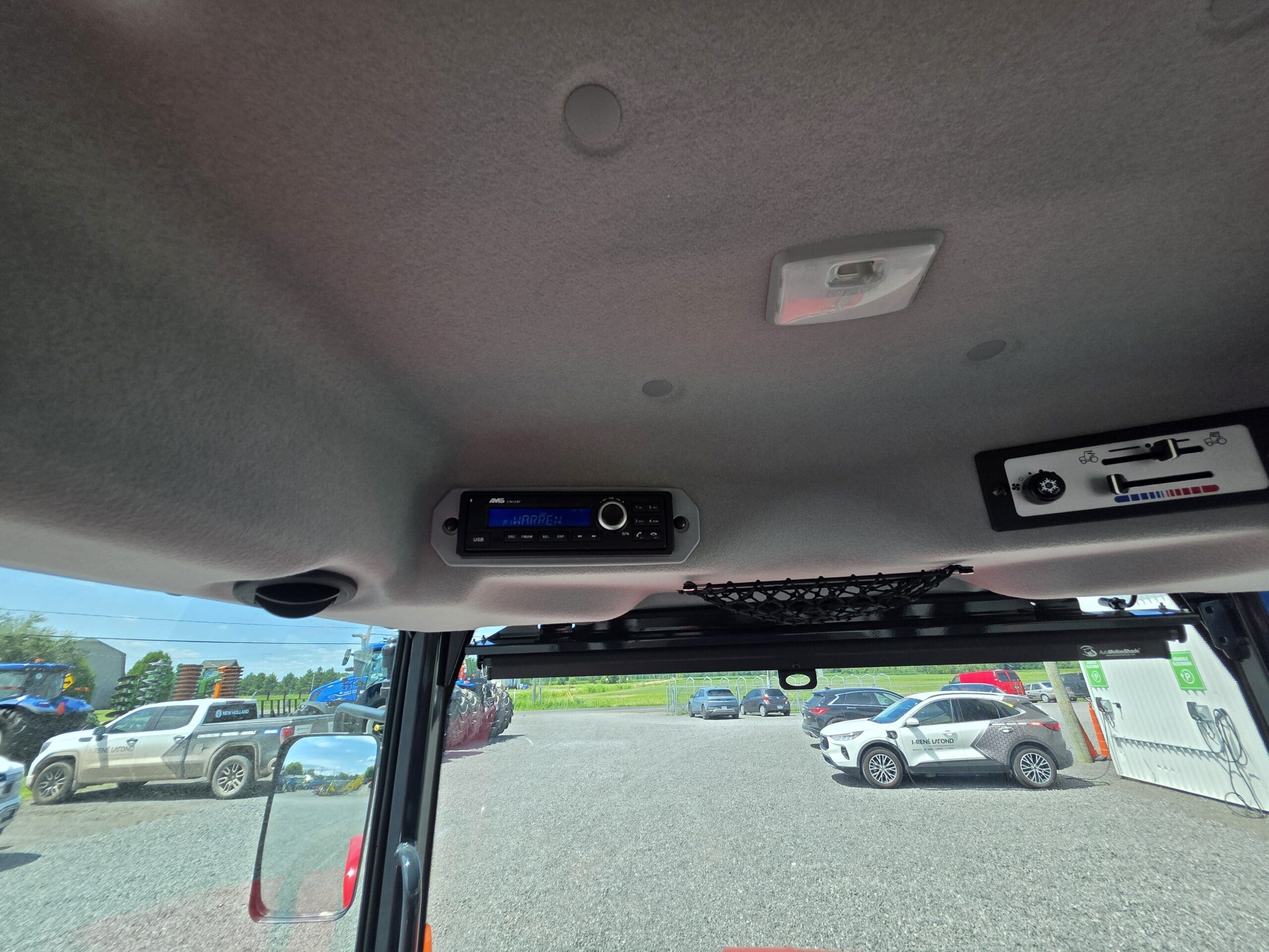Vue de l'intérieur d'un véhicule vers le plafond, montrant une radio et un panneau de commande. Par les fenêtres, on aperçoit des voitures en stationnement et des paysages extérieurs sous un ciel lumineux.