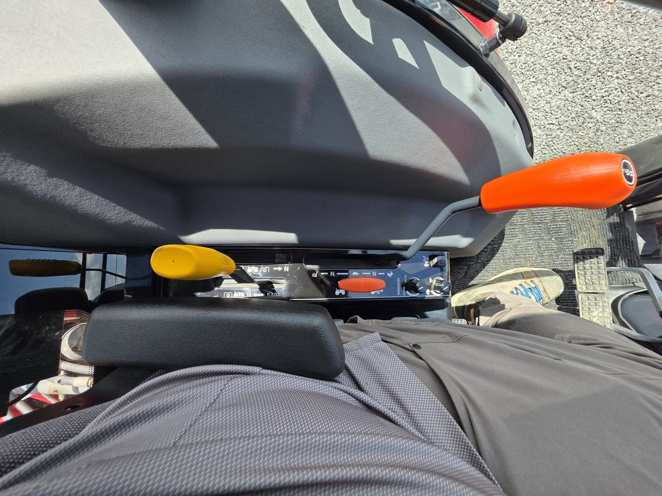 Vue de dessus d'une personne assise sur le siège d'un tracteur, montrant deux leviers de vitesse, l'un jaune, l'autre orange, une partie du sol et les pieds de la personne, chaussée de baskets et vêtue d'un pantalon noir. La lumière du soleil projette des ombres sur le siège.