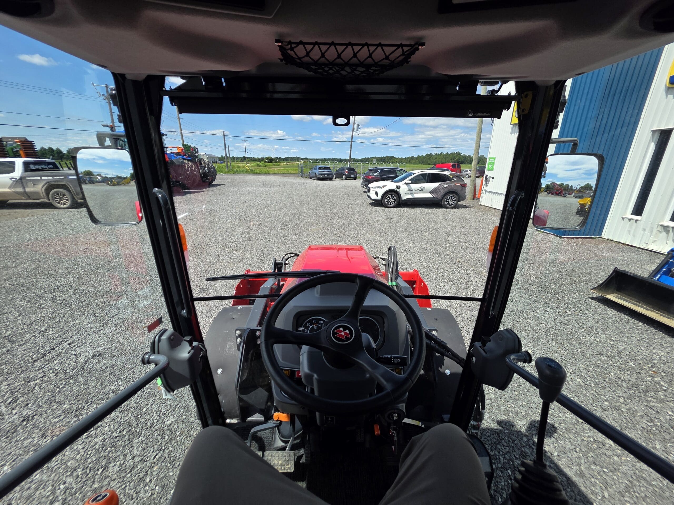 Vue de l'intérieur d'une cabine de tracteur montrant le volant, le tableau de bord et le terrain extérieur avec des véhicules garés et un ciel bleu ensoleillé en arrière-plan.