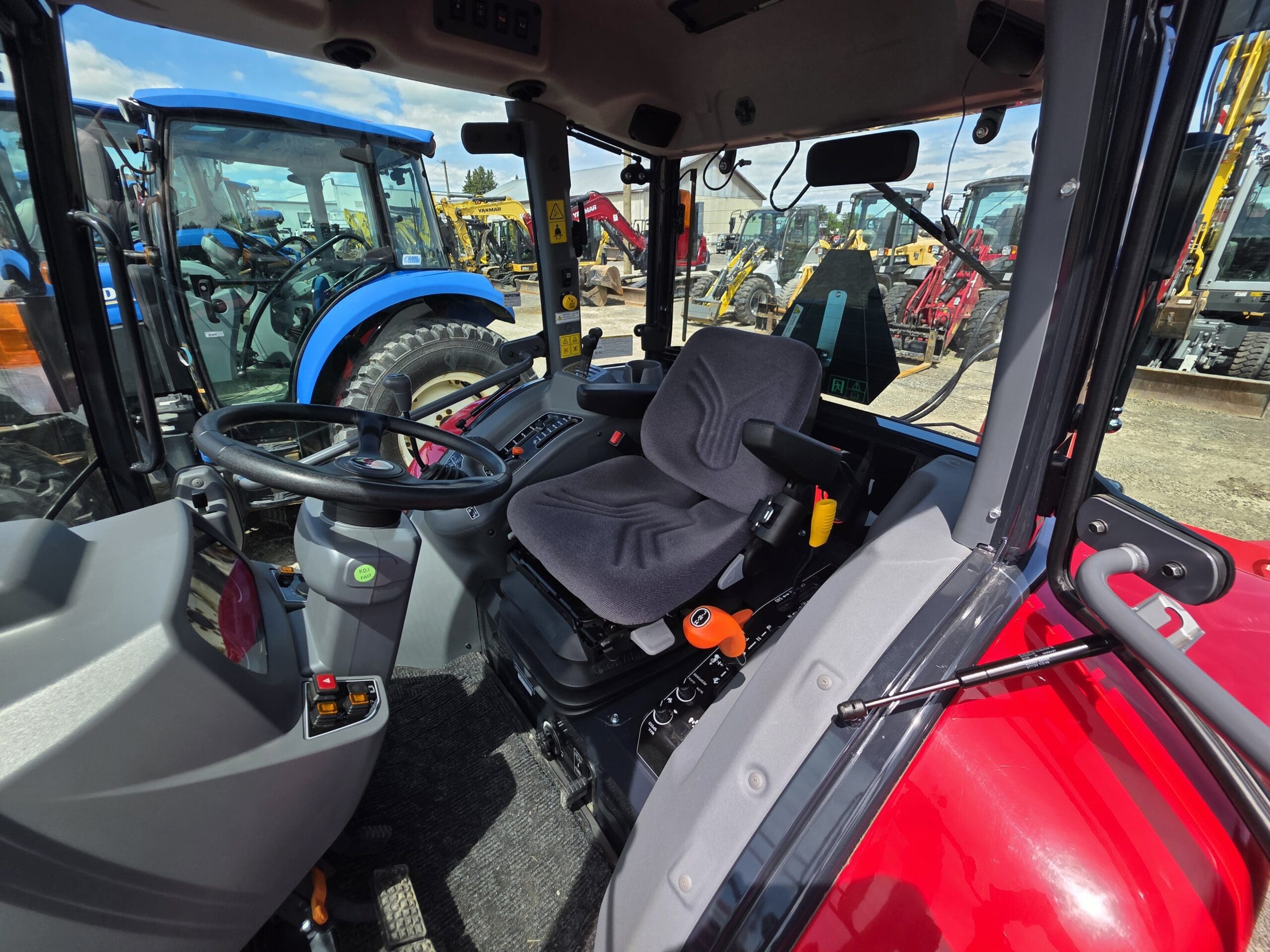 Vue intérieure d'une cabine de tracteur moderne avec un siège noir, un volant, des panneaux de commande et de grandes fenêtres ; d'autres tracteurs et machines sont visibles à l'extérieur dans un terrain sous un ciel bleu.
