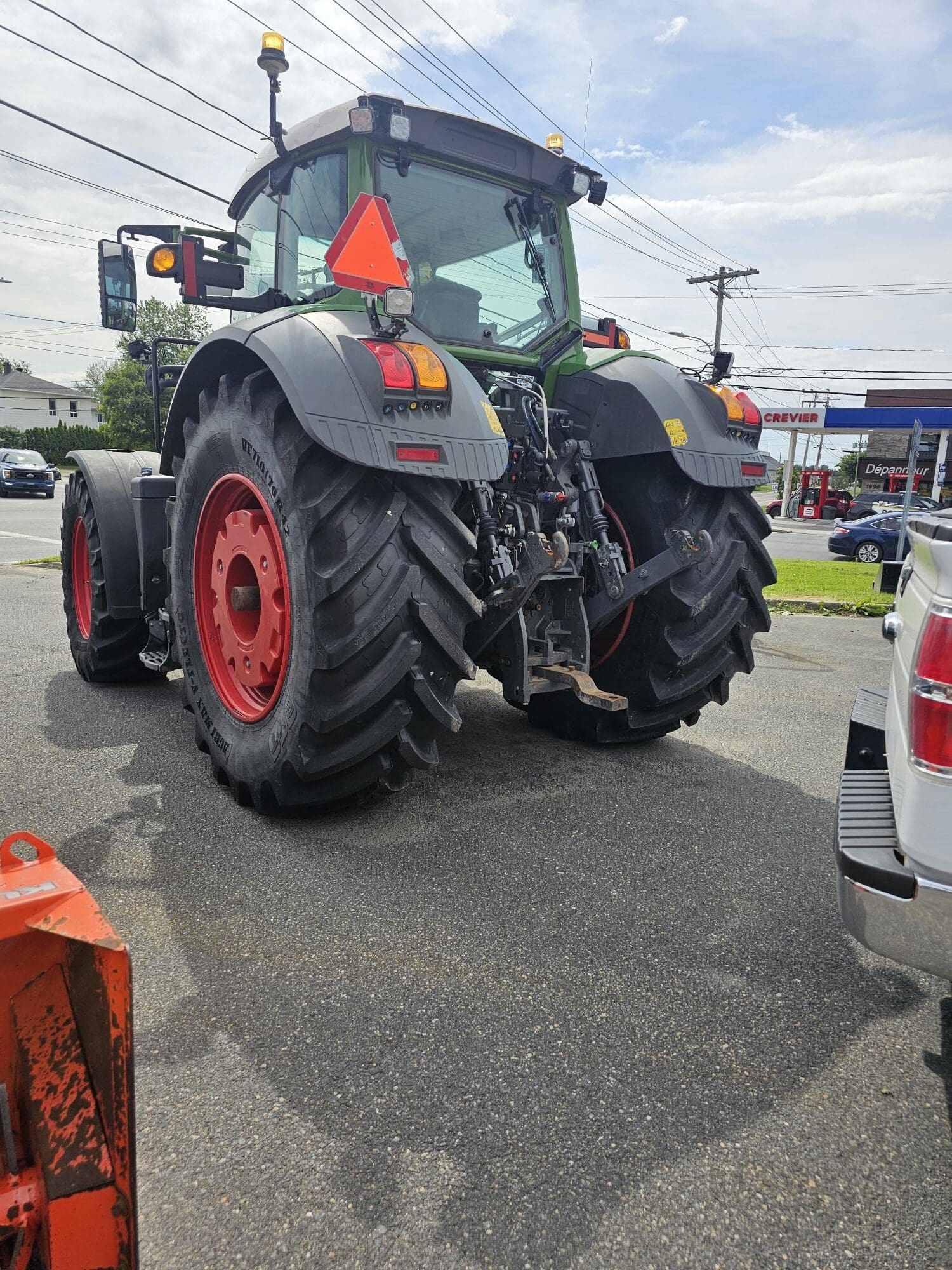 Un grand tracteur moderne avec des pneus arrière surdimensionnés est garé sur la chaussée à côté d'un véhicule blanc. Des lignes électriques, des bâtiments et quelques voitures sont visibles à l'arrière-plan sous un ciel partiellement nuageux.