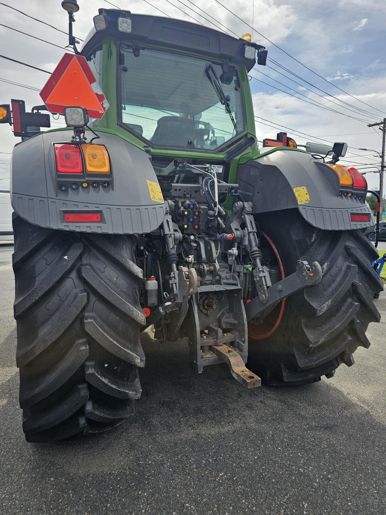 Un tracteur vert vu de l'arrière, montrant de gros pneus arrière noirs, diverses pièces mécaniques, des feux arrière et un triangle réfléchissant orange vif pour la sécurité. Le tracteur est garé sur l'asphalte sous un ciel partiellement nuageux.