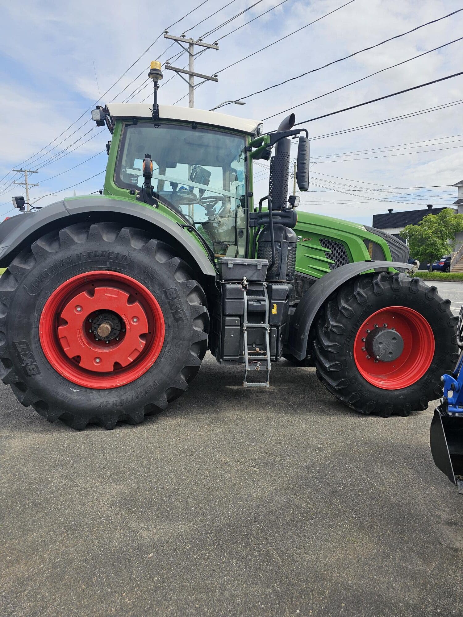 Un grand tracteur vert avec des pneus noirs et des jantes rouges est garé sur une surface pavée sous un ciel partiellement nuageux, avec des poteaux et des lignes électriques visibles à l'arrière-plan.