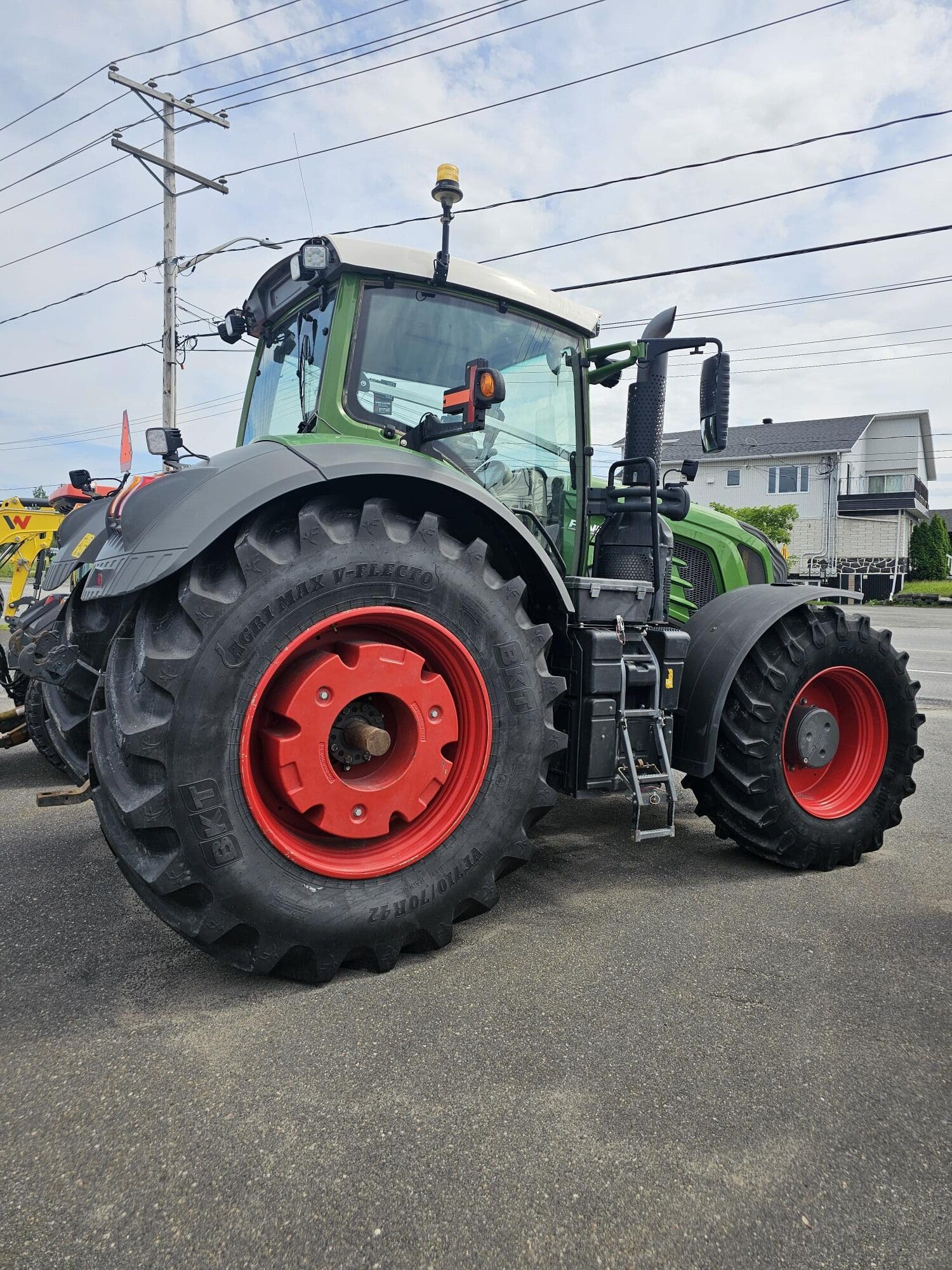 Un tracteur vert avec de gros pneus noirs et des jantes rouges est garé sur une surface pavée près d'une zone résidentielle avec des maisons et des lignes électriques en arrière-plan.