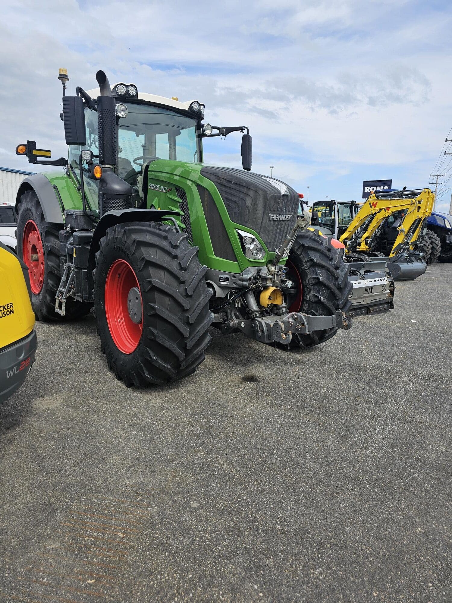 Un grand tracteur Fendt vert avec des roues rouges est garé en plein air sur un trottoir, entouré d'autres machines de construction et agricoles, sous un ciel plutôt nuageux.