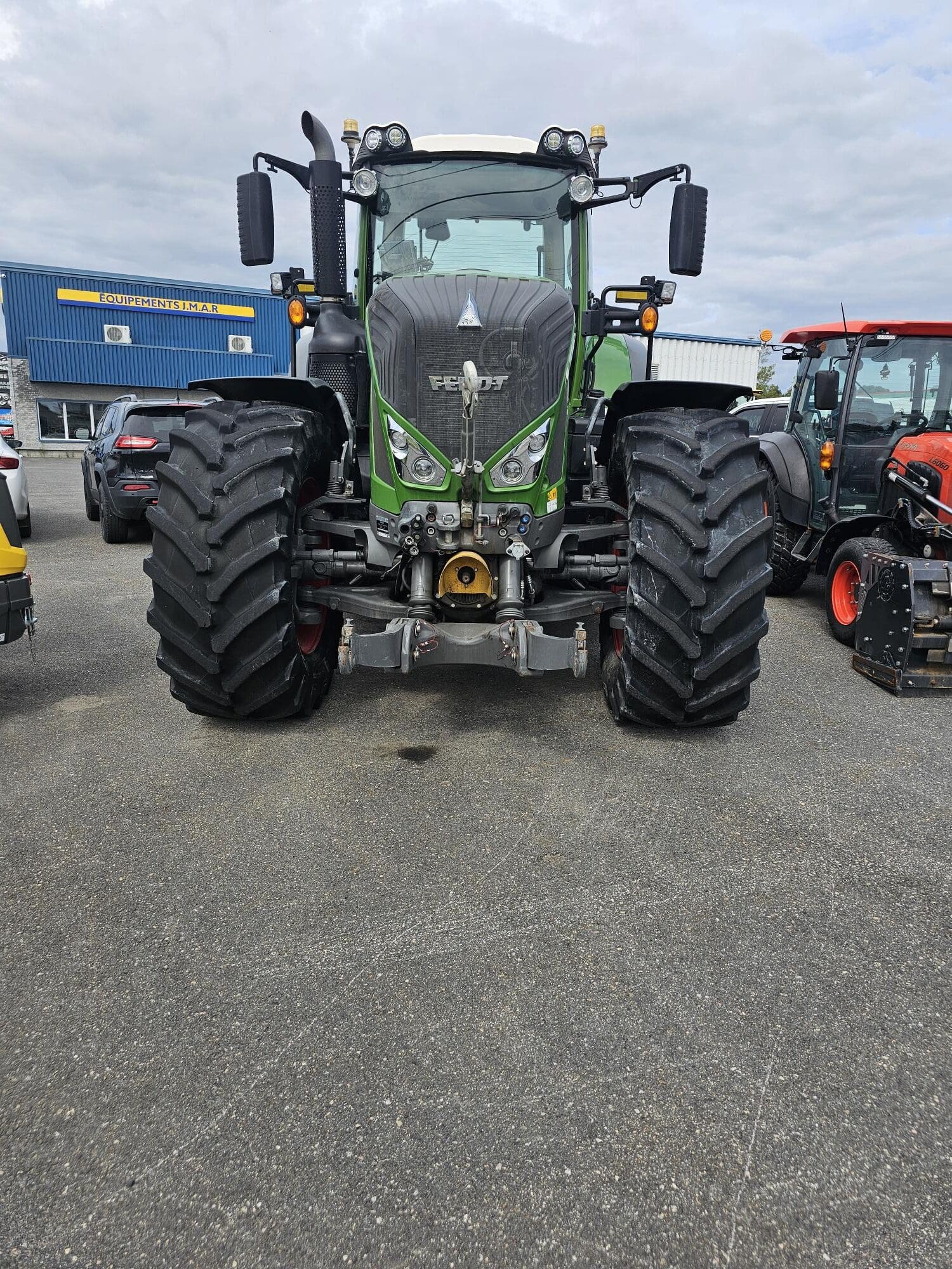 Un grand tracteur Fendt vert aux pneus massifs est garé sur une surface asphaltée. D'autres véhicules et un bâtiment industriel bleu sont visibles à l'arrière-plan sous un ciel nuageux.