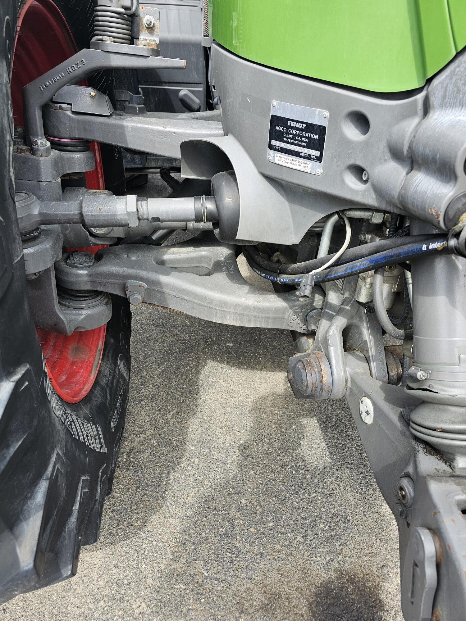 Vue rapprochée de la roue avant et du système de suspension d'un tracteur, montrant des pièces mécaniques détaillées, des conduites hydrauliques et une étiquette du fabricant sur un panneau de carrosserie vert. Le sol est recouvert d'asphalte.
