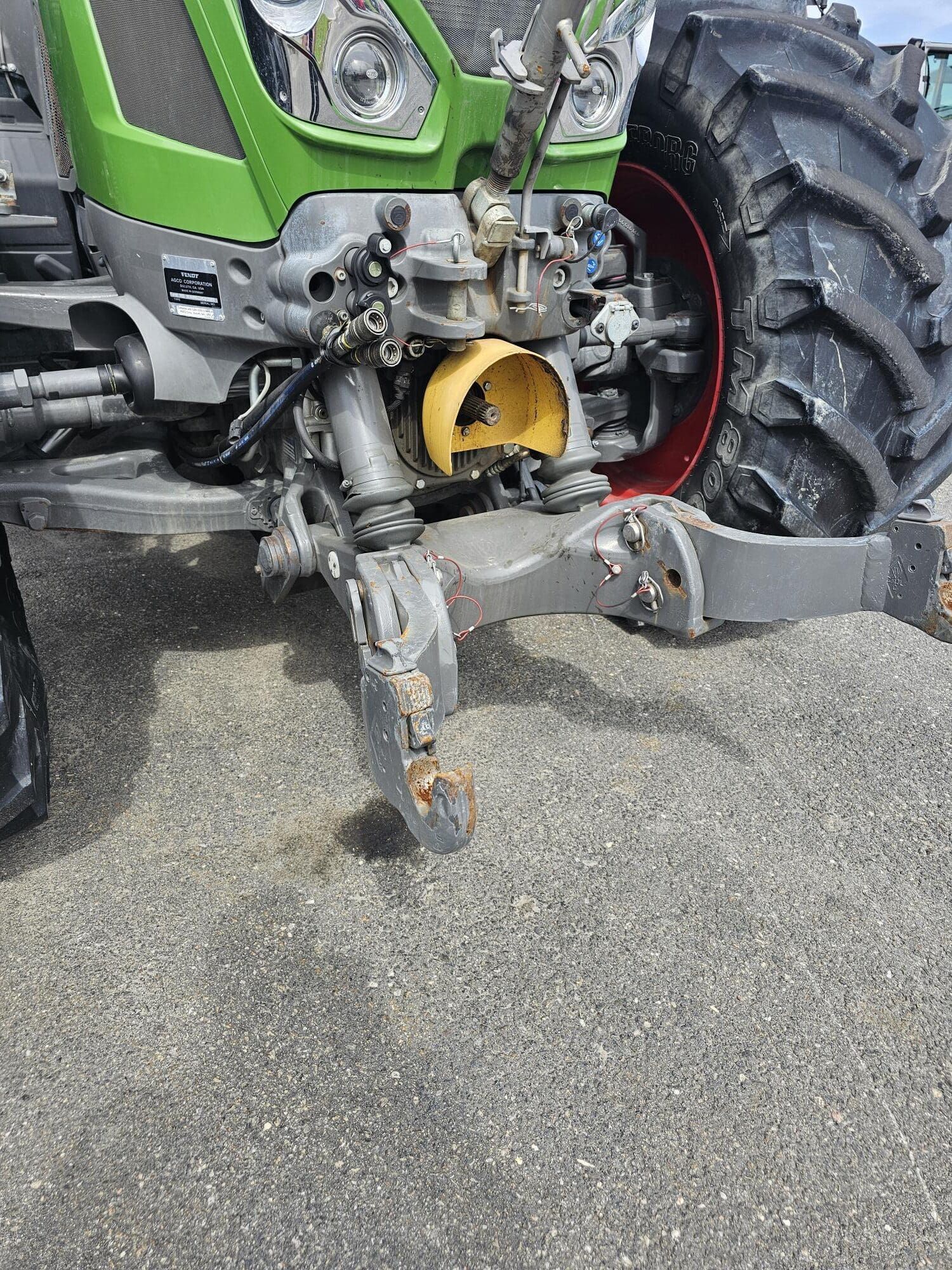 Gros plan de l'avant d'un tracteur vert, montrant ses connexions hydrauliques, un couvercle jaune d'arbre à cardans et un attelage avant rouillé sur une surface pavée.