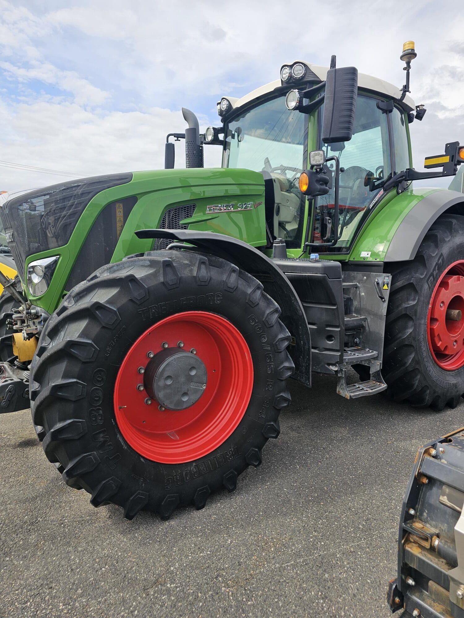 Un grand tracteur vert aux roues rouge vif est garé sur une surface pavée sous un ciel partiellement nuageux. Le tracteur est équipé de pneus résistants et d'équipements modernes.