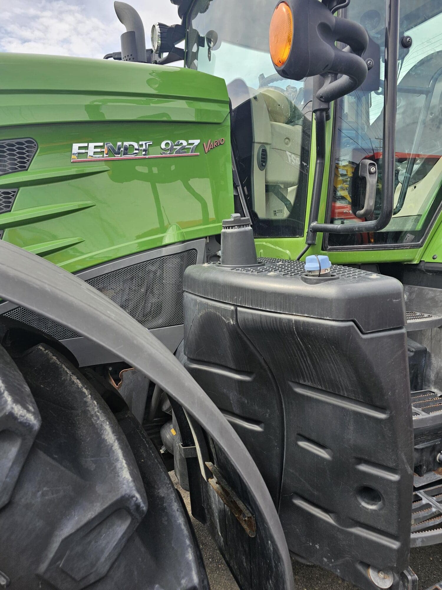 Gros plan d'un tracteur Fendt 927 Vario vert, montrant la roue avant, le réservoir de carburant et une partie de la cabine avec un voyant orange visible.