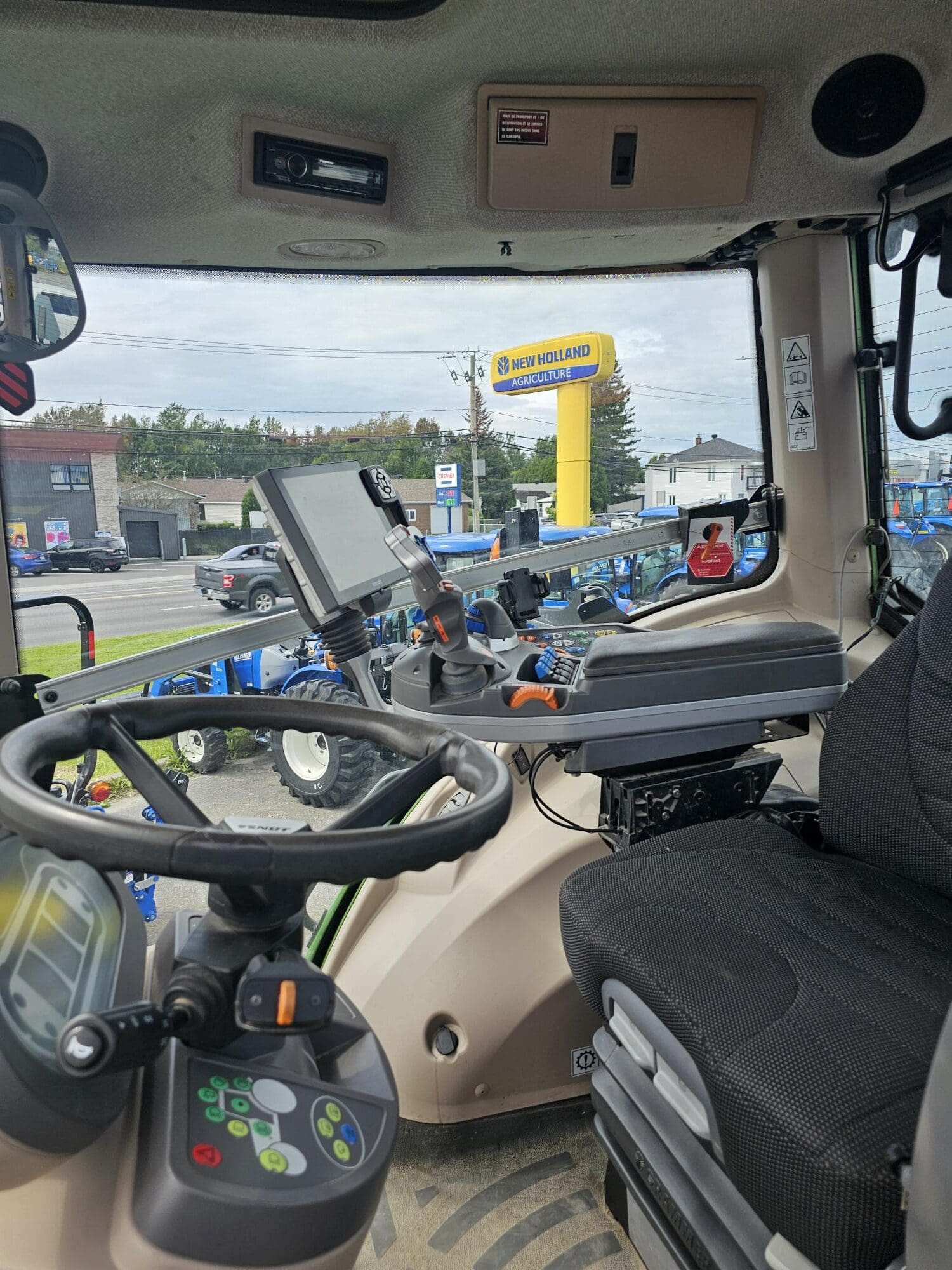 Vue de l'intérieur d'une cabine de tracteur moderne montrant le siège du conducteur, les panneaux de commande, l'affichage numérique et le volant, avec un concessionnaire New Holland et des tracteurs visibles à l'extérieur par la fenêtre.