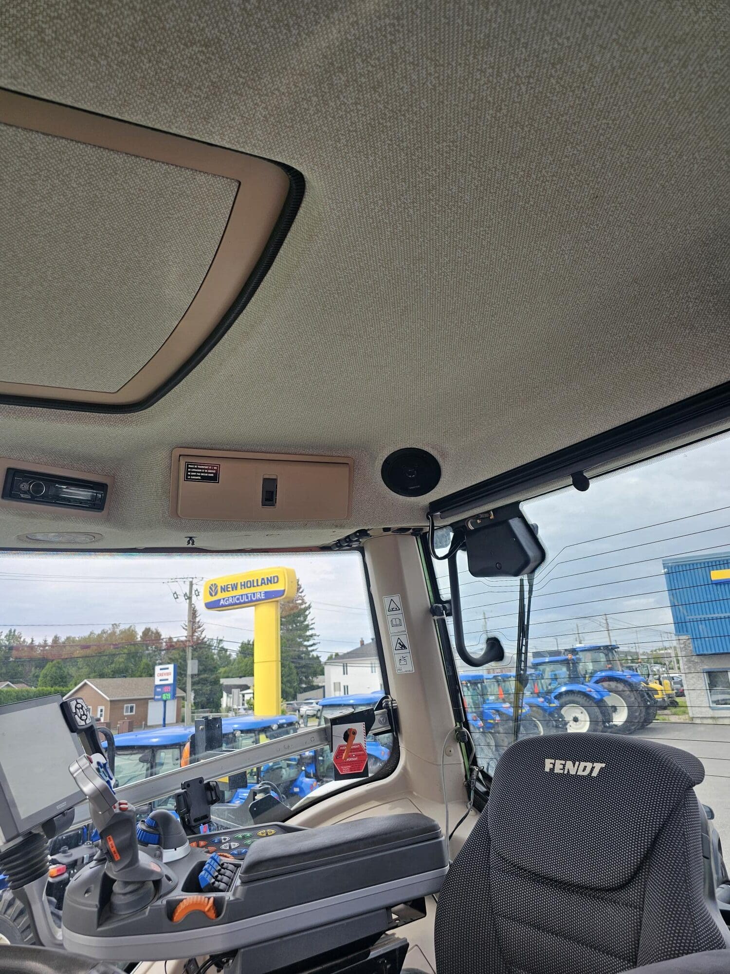 Vue de l'intérieur de la cabine d'un tracteur Fendt, montrant le siège du conducteur, le panneau de commande et une partie du plafond. Par les fenêtres, on aperçoit à l'extérieur des tracteurs New Holland et un panneau jaune New Holland.