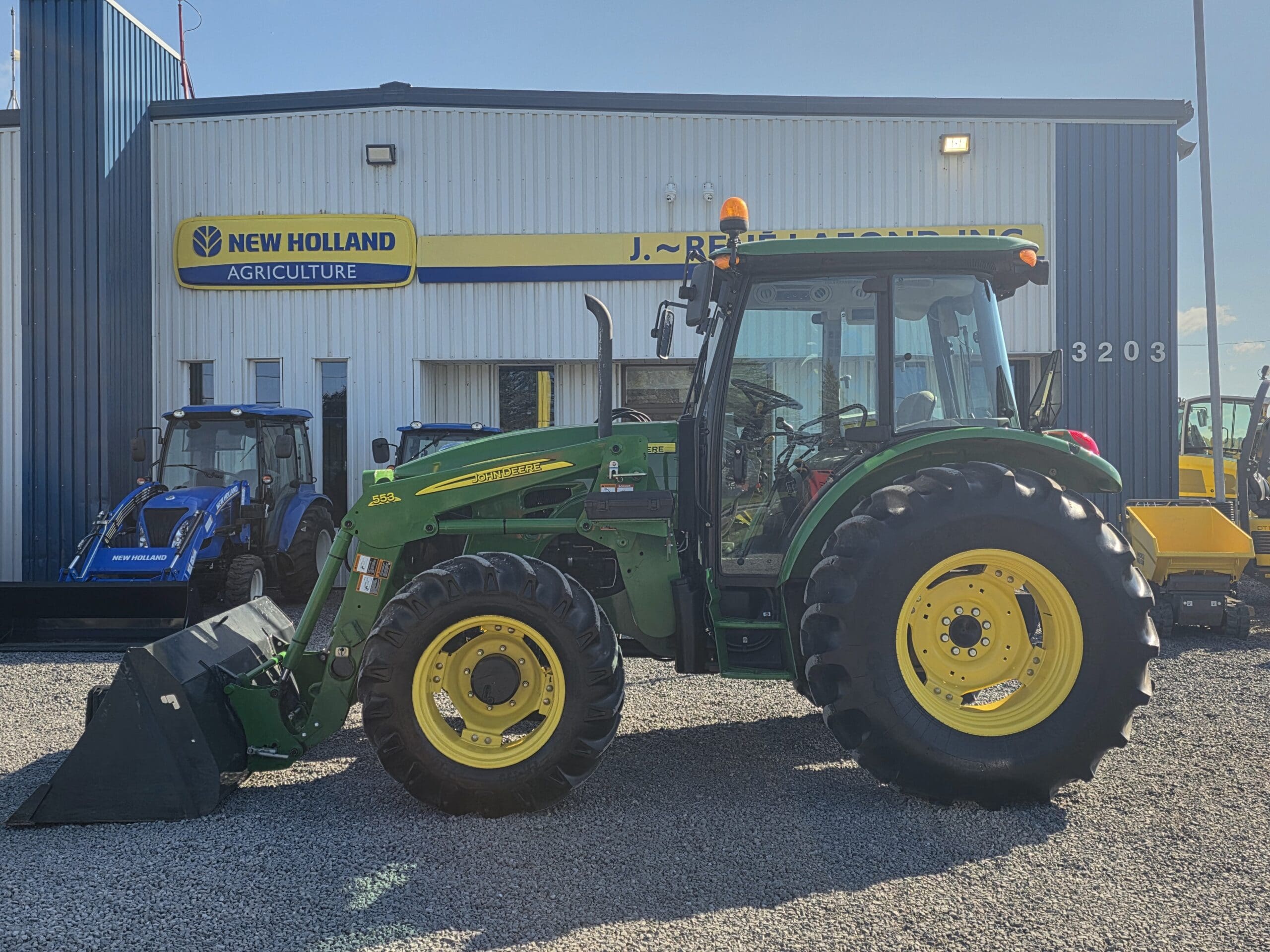 Un tracteur John Deere vert et jaune, équipé d'un chargeur frontal, est garé devant le bâtiment d'un concessionnaire New Holland Agriculture par une journée ensoleillée. Un autre tracteur bleu est visible à l'arrière-plan.