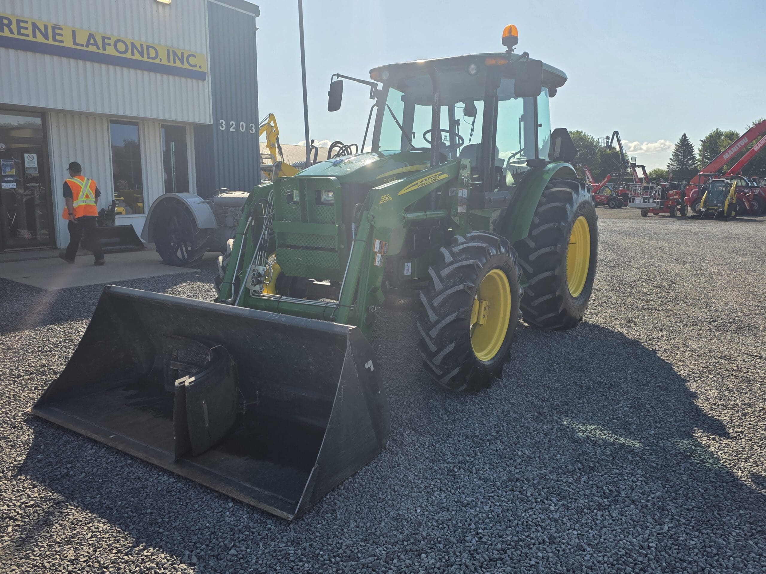 Un tracteur vert et jaune avec un chargeur frontal est garé sur du gravier devant un bâtiment. Deux travailleurs portant des gilets de sécurité se trouvent à proximité, et d'autres machines sont visibles à l'arrière-plan.