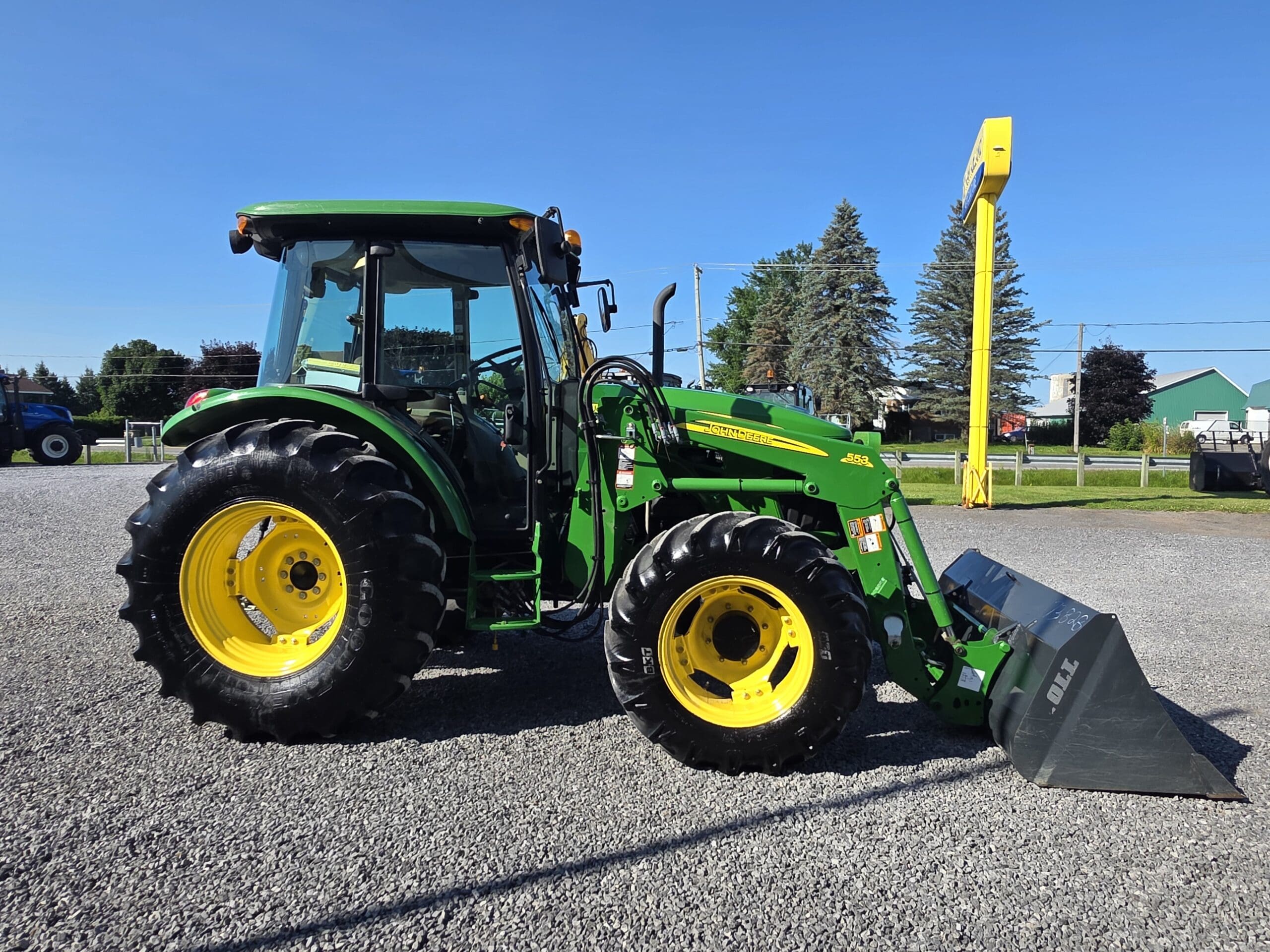 Un tracteur John Deere vert avec de grandes roues jaunes et un chargeur frontal est garé sur une surface de gravier sous un ciel bleu clair. Des arbres et un panneau jaune sont visibles à l'arrière-plan.