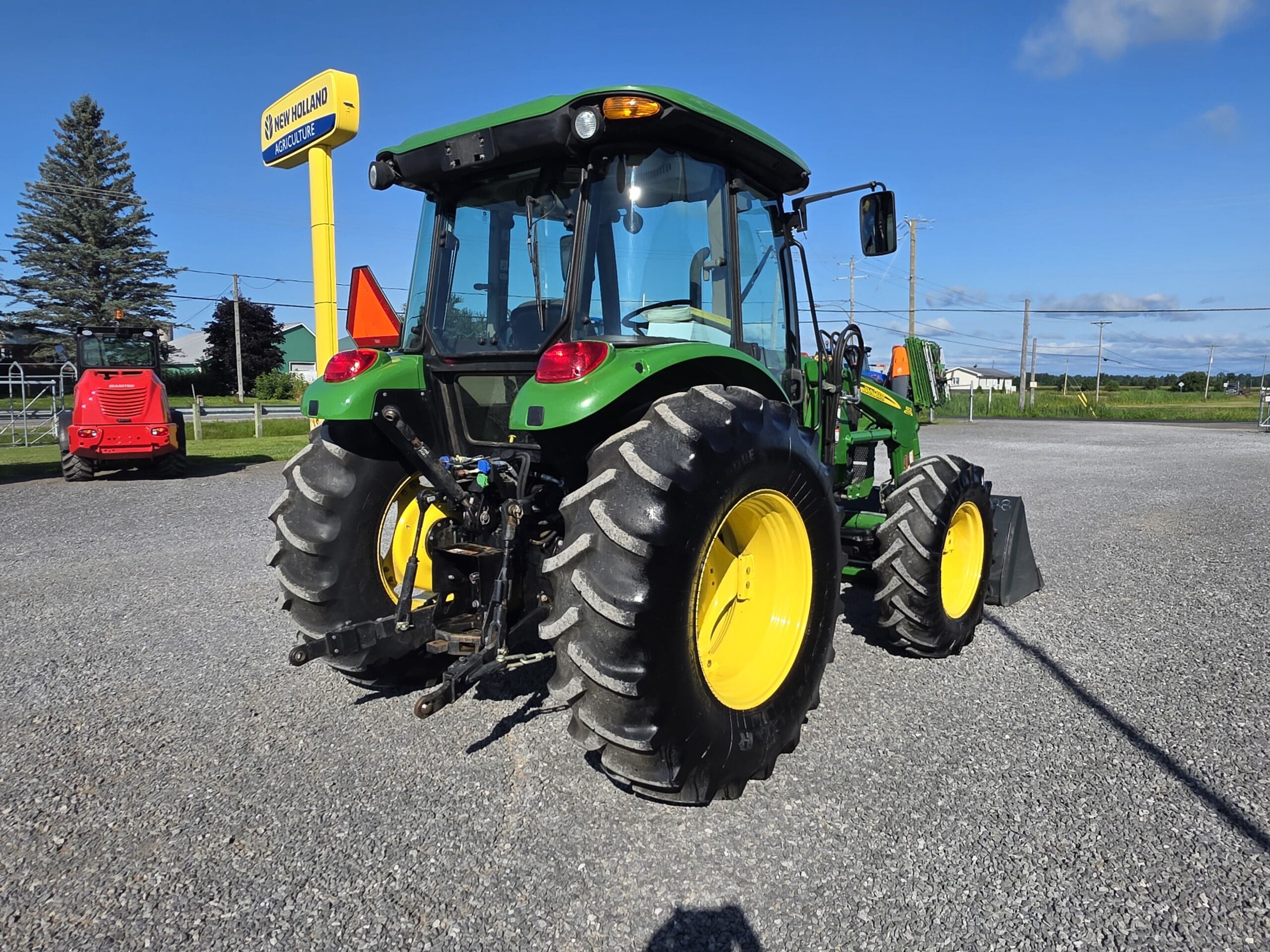 Un tracteur John Deere vert et jaune est garé sur du gravier près de l'enseigne d'un concessionnaire New Holland, avec un ciel bleu clair et des champs herbeux en arrière-plan.