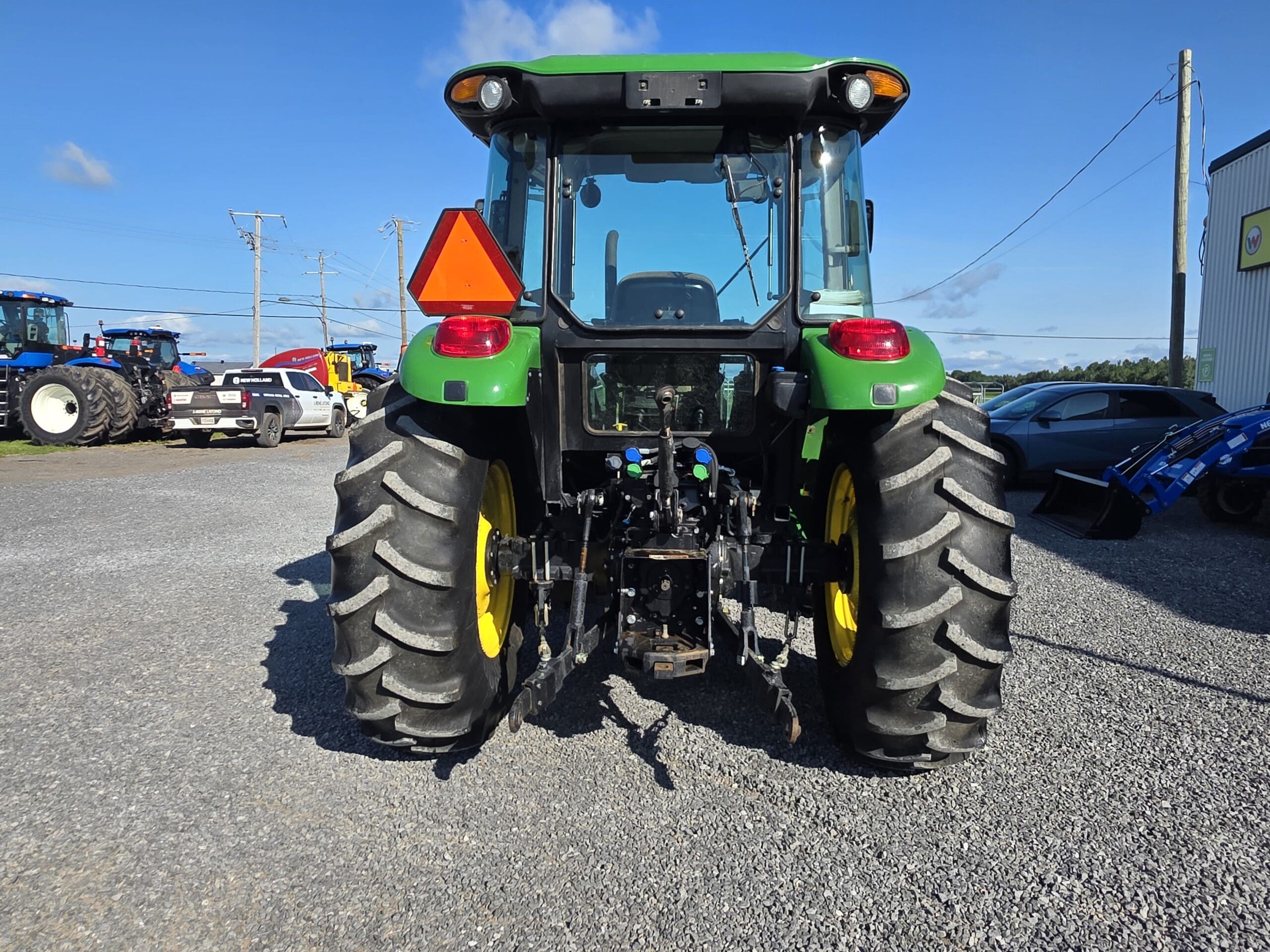Vue arrière d'un tracteur vert garé sur du gravier, montrant de gros pneus noirs avec des jantes jaunes, un panneau de sécurité triangulaire orange et du matériel agricole attaché à l'arrière.