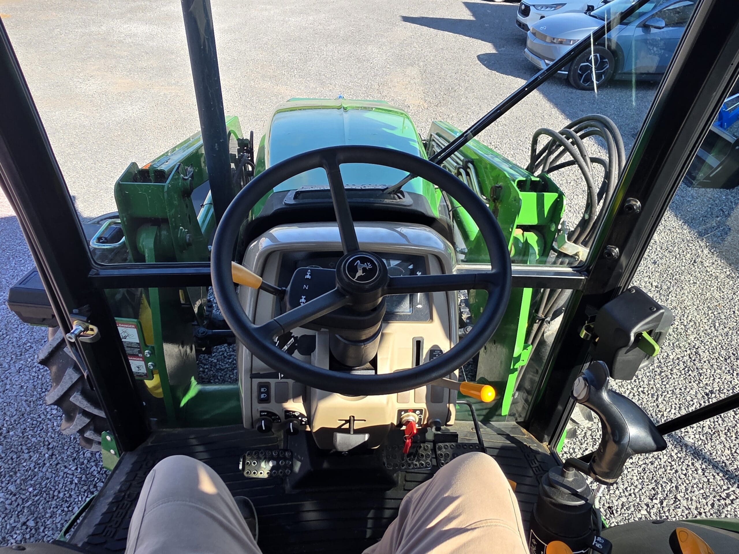 Vue du siège du conducteur d'un tracteur vert, montrant le volant, les commandes du tableau de bord, les pédales et un aperçu des jambes du conducteur. Plusieurs véhicules sont garés à l'extérieur sur une surface en gravier.