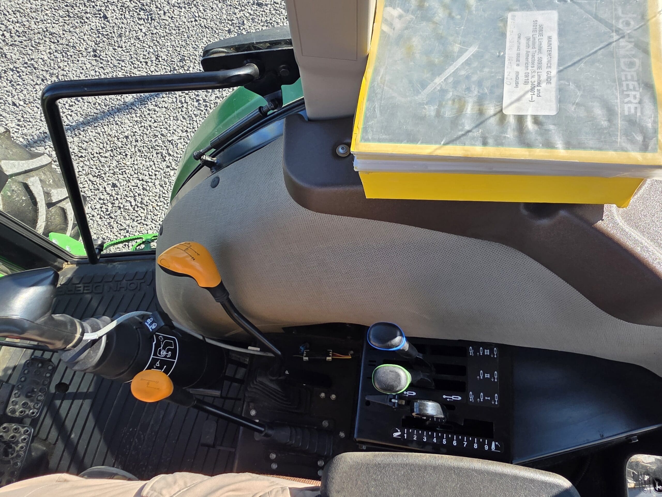 Vue de l'intérieur d'un tracteur, montrant le siège, les leviers de vitesse, le panneau de commande, un manuel John Deere dans un classeur jaune et une partie du volant. La lumière du soleil projette des ombres sur la zone.