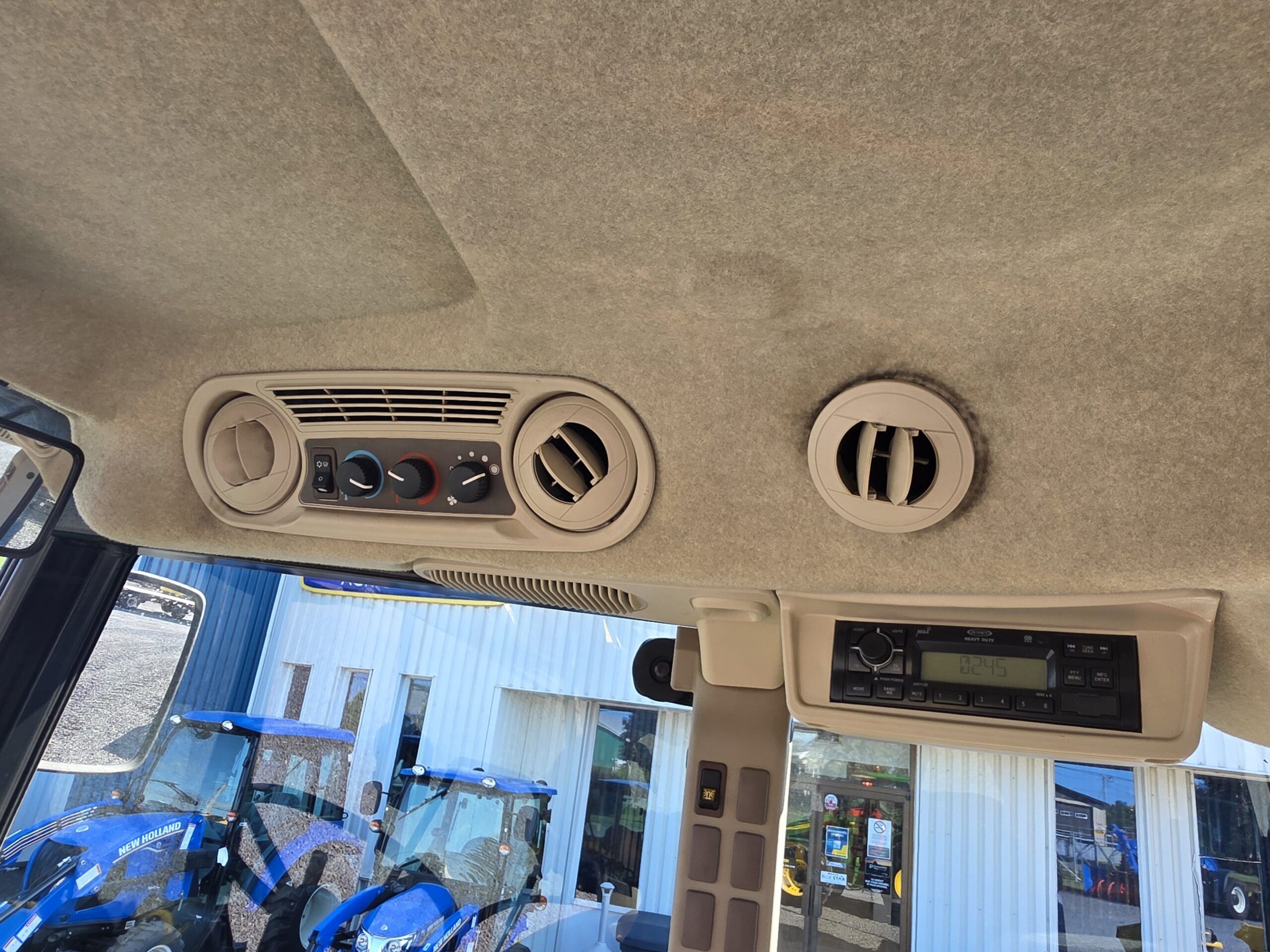 L'image montre le panneau de toit intérieur d'un véhicule, avec des bouches d'air conditionné, des boutons de réglage de la climatisation et un système radio. Par la fenêtre, on aperçoit des tracteurs bleus et un bâtiment à l'extérieur.