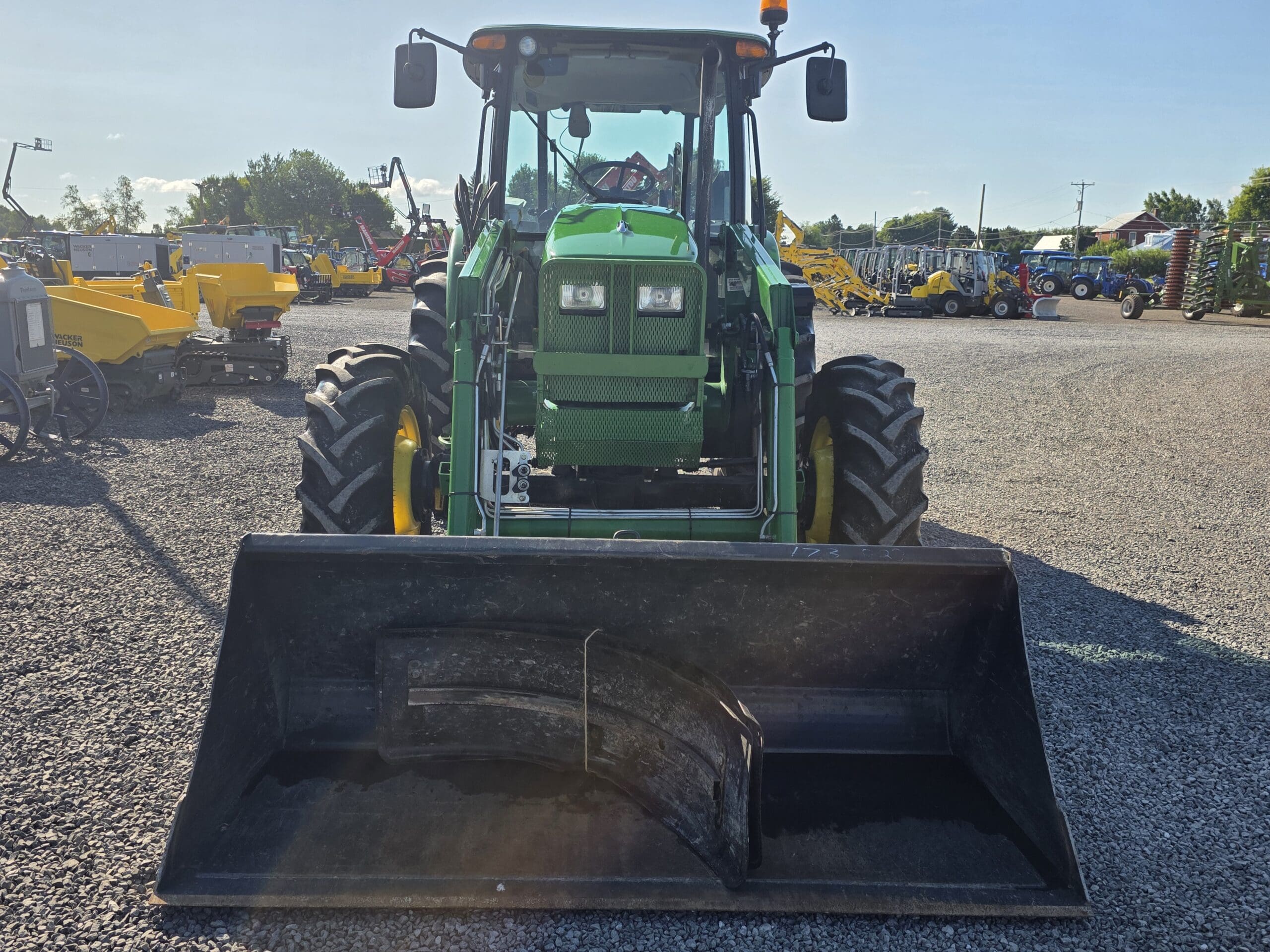 Un tracteur vert équipé d'un chargeur frontal est garé sur un terrain en gravier, entouré de divers autres équipements agricoles jaunes et bleus, sous un ciel ensoleillé.