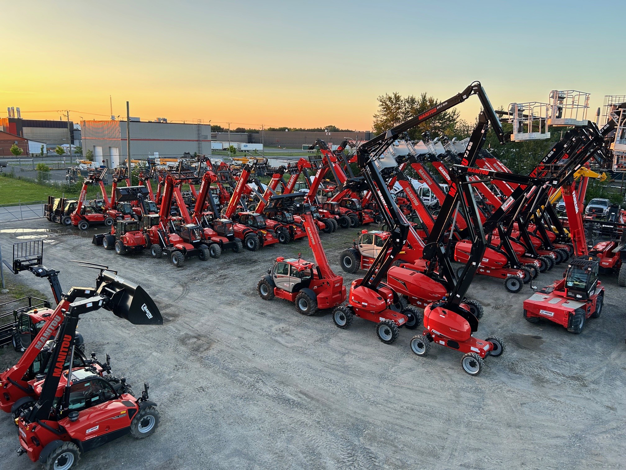 Une grande cour remplie de nombreux véhicules de construction rouges, y compris des télescopiques et des nacelles élévatrices New Holland, garés en rangées sur du gravier au coucher du soleil, avec des bâtiments industriels et l'enseigne Machinerie Avantis St-Vallier à l'arrière-plan.