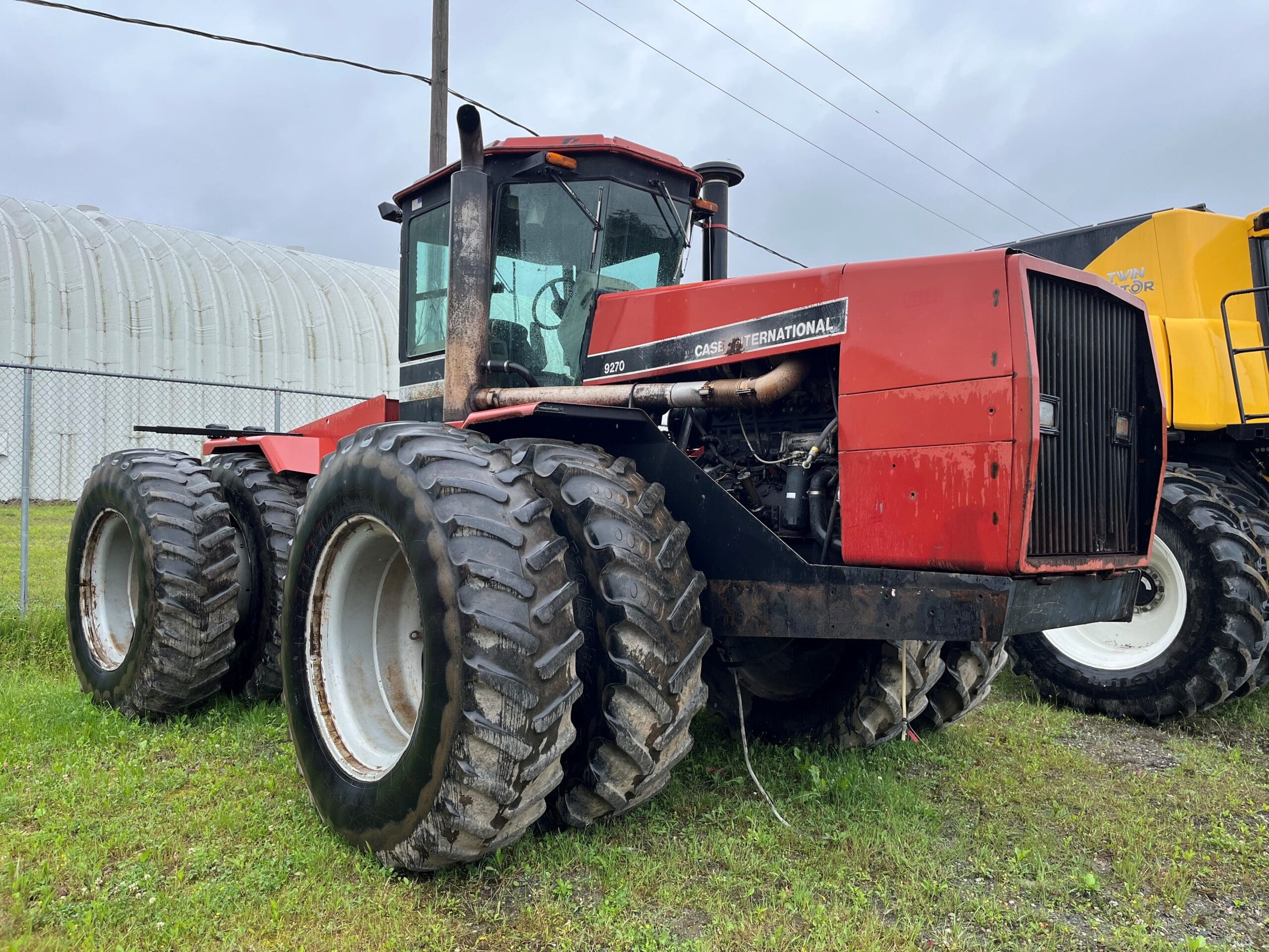 Un gros tracteur Case International rouge avec six gros pneus est garé sur l'herbe près d'une clôture métallique et d'une grande structure de stockage, sous un ciel nuageux.