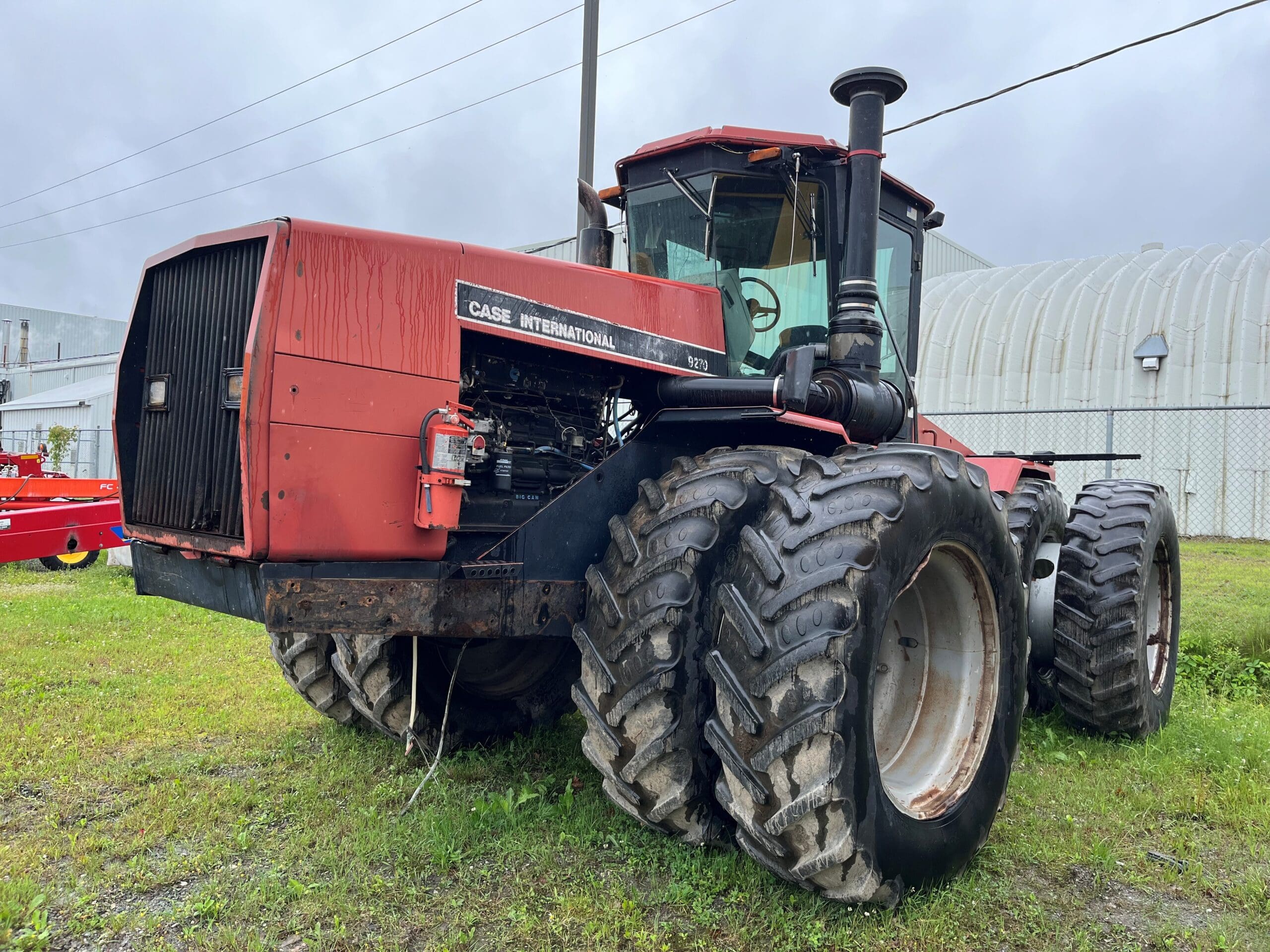Un tracteur Case International 9170 rouge avec de grandes roues arrière doubles est garé sur l'herbe près d'un bâtiment industriel blanc et d'un ciel nuageux. Le tracteur semble usé et usé.