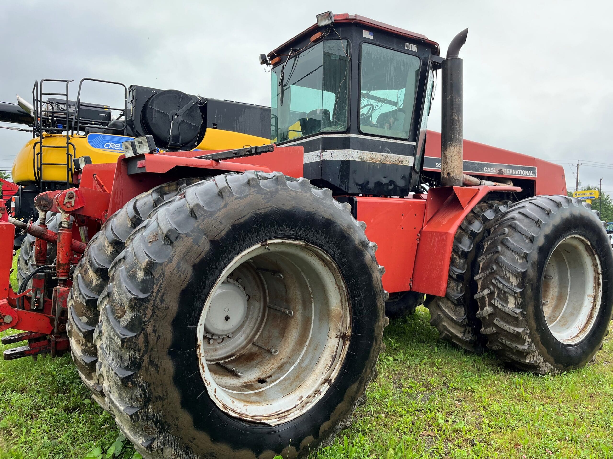 Un grand tracteur Case International rouge avec des roues arrière doubles surdimensionnées est garé sur de l'herbe sous un ciel nuageux. Du matériel agricole est visible à l'arrière-plan.