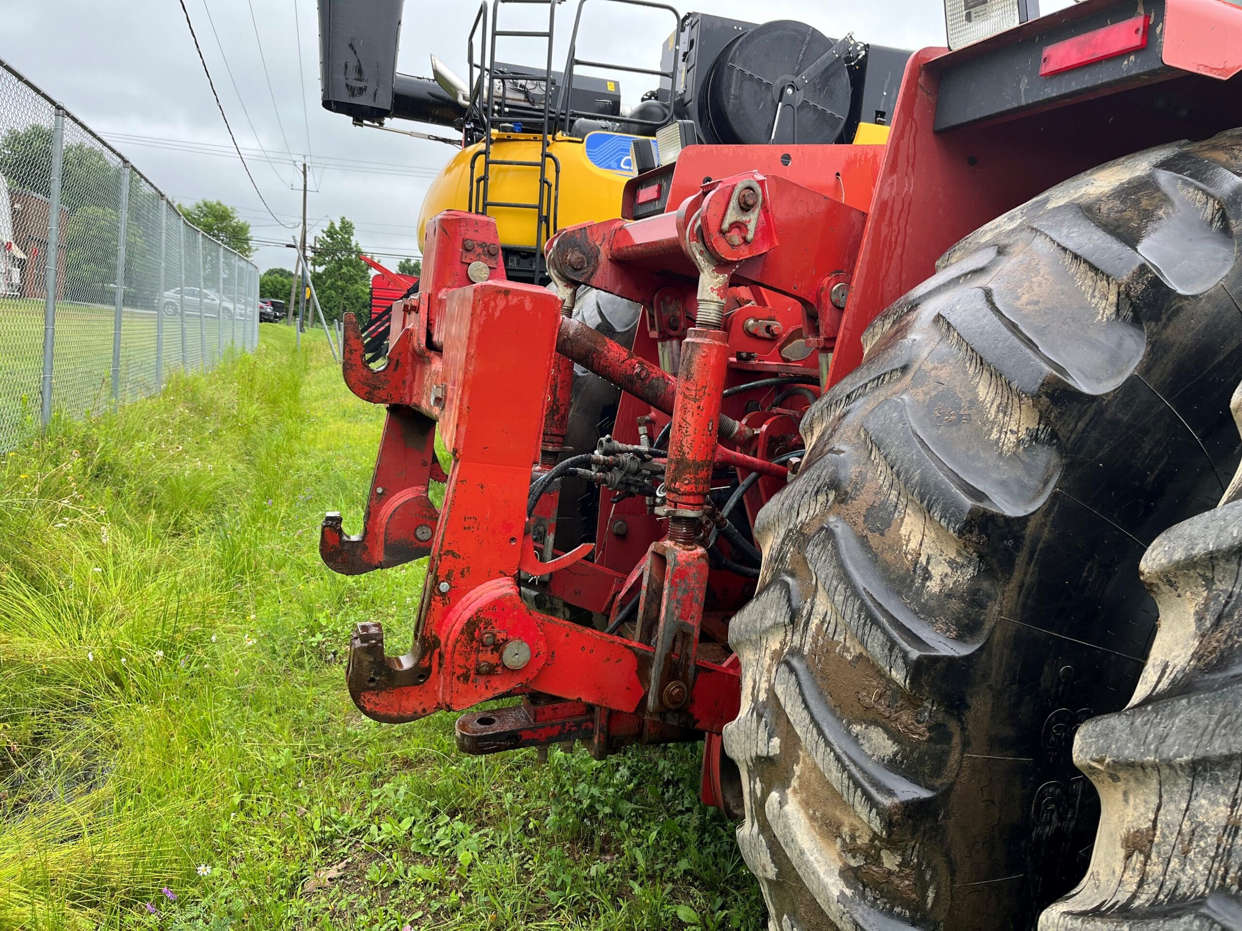 Gros plan de la partie arrière d'un tracteur rouge montrant son gros pneu sale et son mécanisme d'attelage, garé sur l'herbe à côté d'une clôture à mailles losangées, avec une autre machine jaune visible à l'arrière-plan.