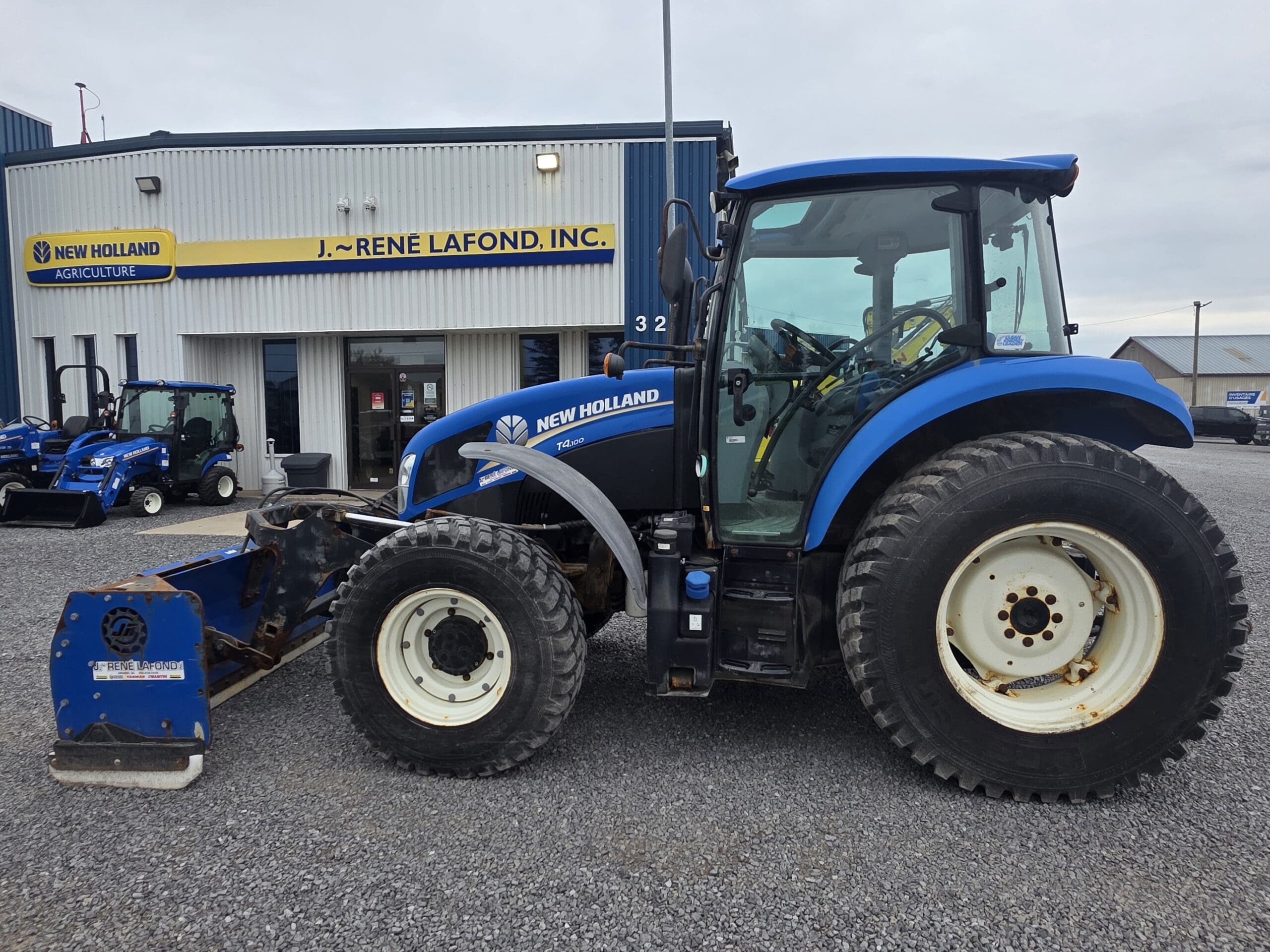 Un tracteur New Holland bleu à gros pneus est garé sur du gravier devant un bâtiment portant l'inscription