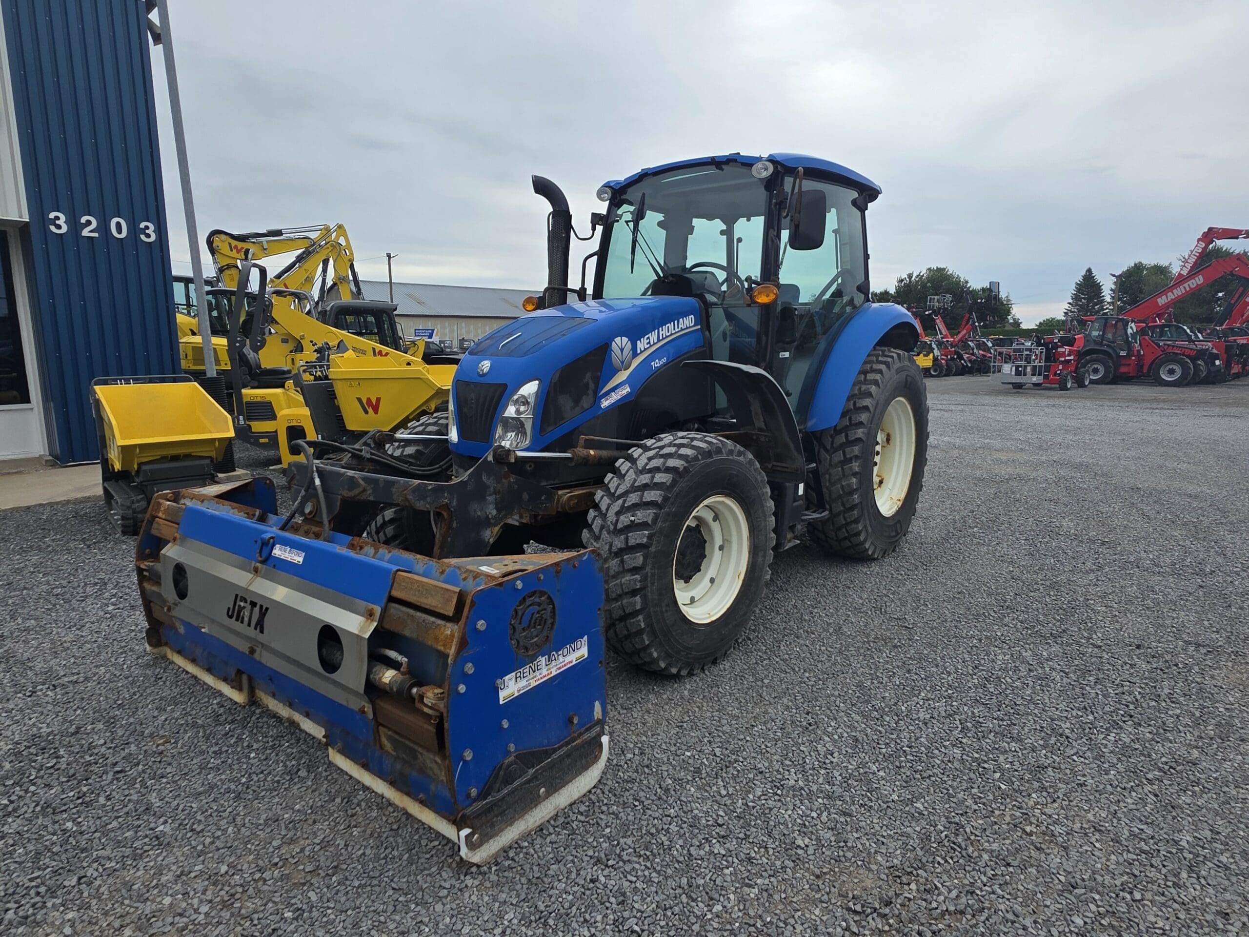 Un tracteur New Holland bleu équipé d'un accessoire JETX est garé sur du gravier à l'extérieur d'un bâtiment. D'autres machines jaunes et rouges sont visibles à l'arrière-plan.