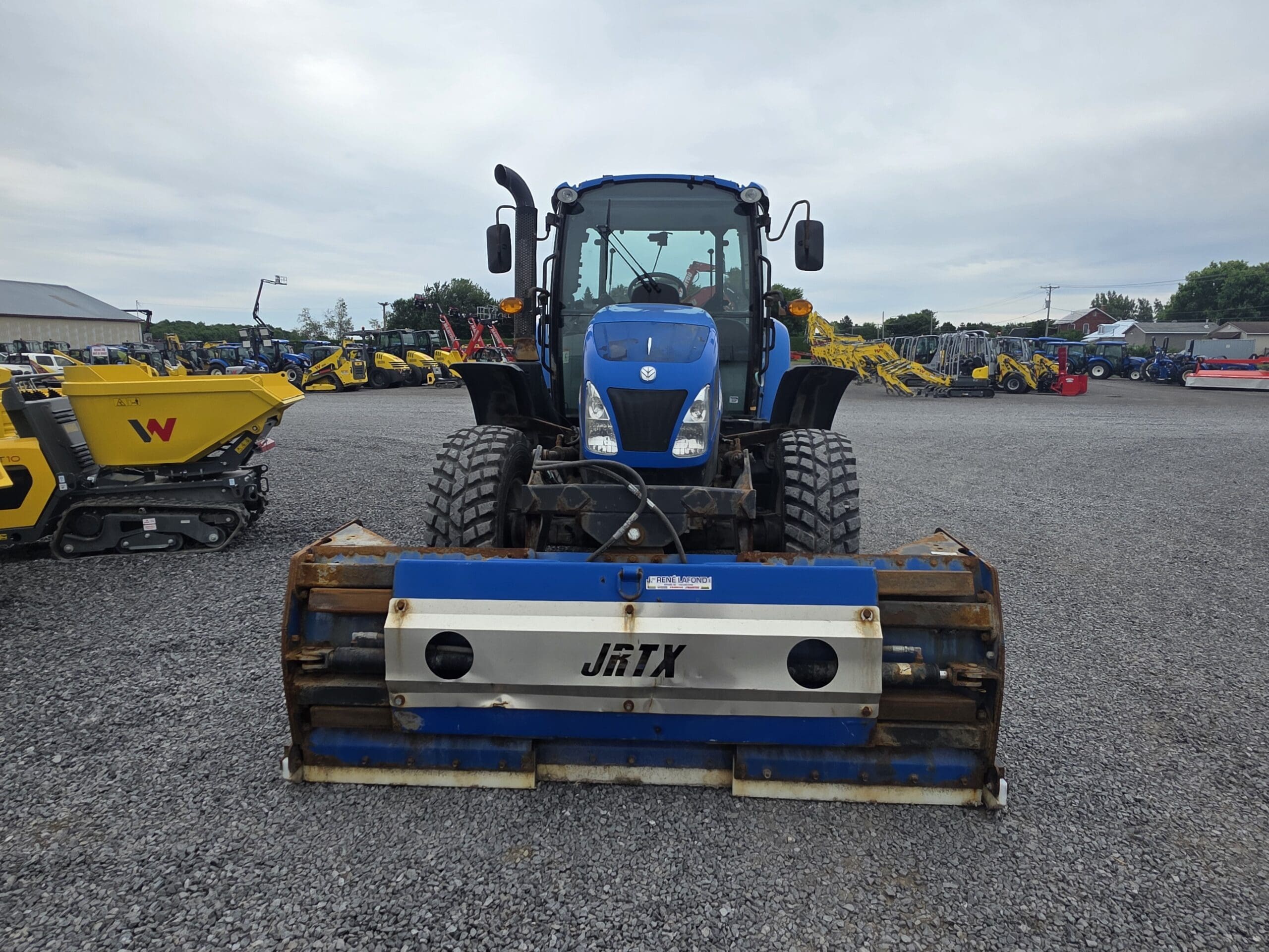 Un tracteur bleu équipé d'un accessoire JRTX est garé sur du gravier dans un parc de machines, entouré de divers autres équipements de construction sous un ciel nuageux.
