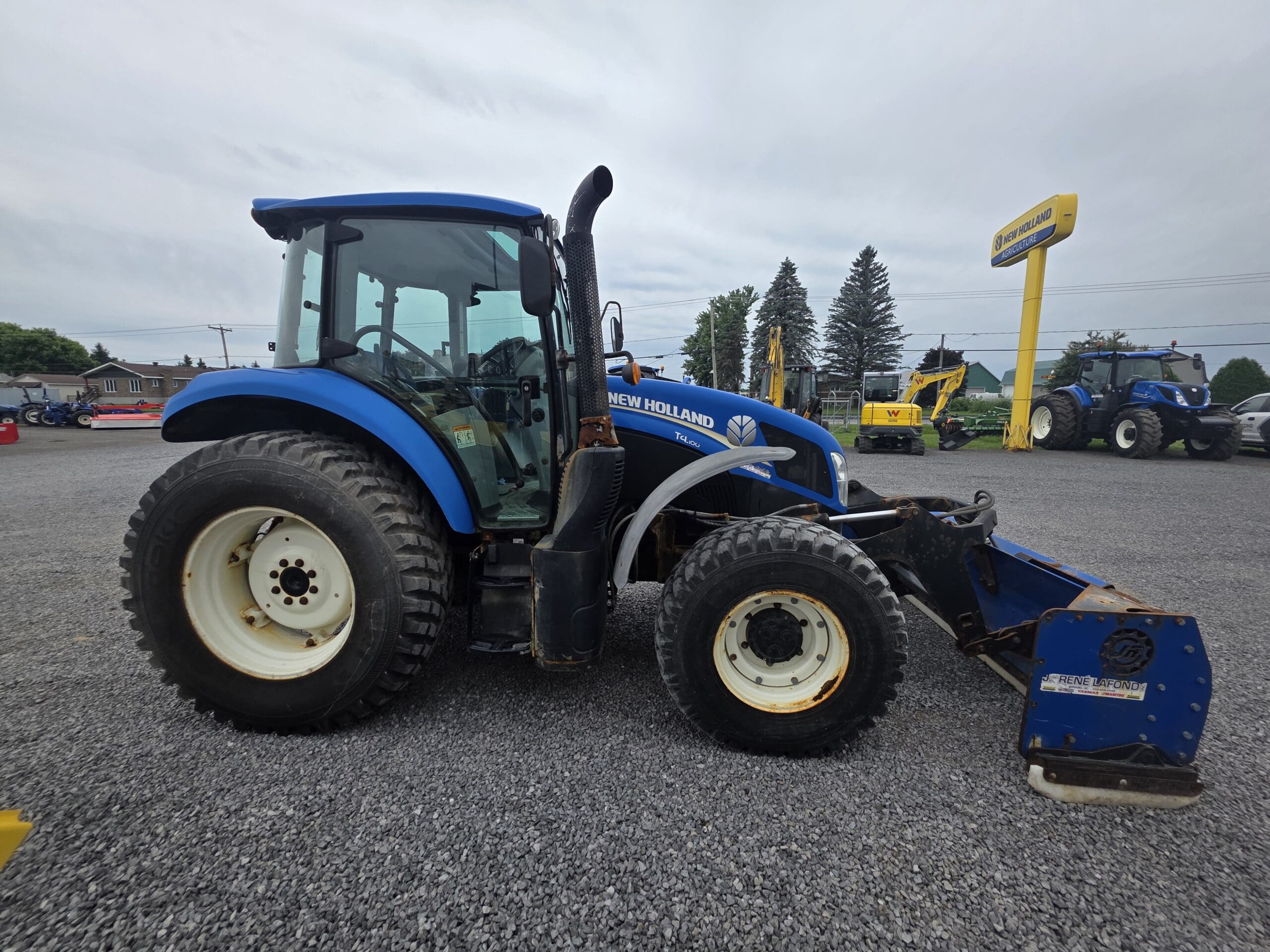 Un tracteur New Holland bleu équipé de gros pneus et d'un outil frontal est garé sur un terrain en gravier. D'autres tracteurs et équipements sont visibles à l'arrière-plan sous un ciel nuageux.