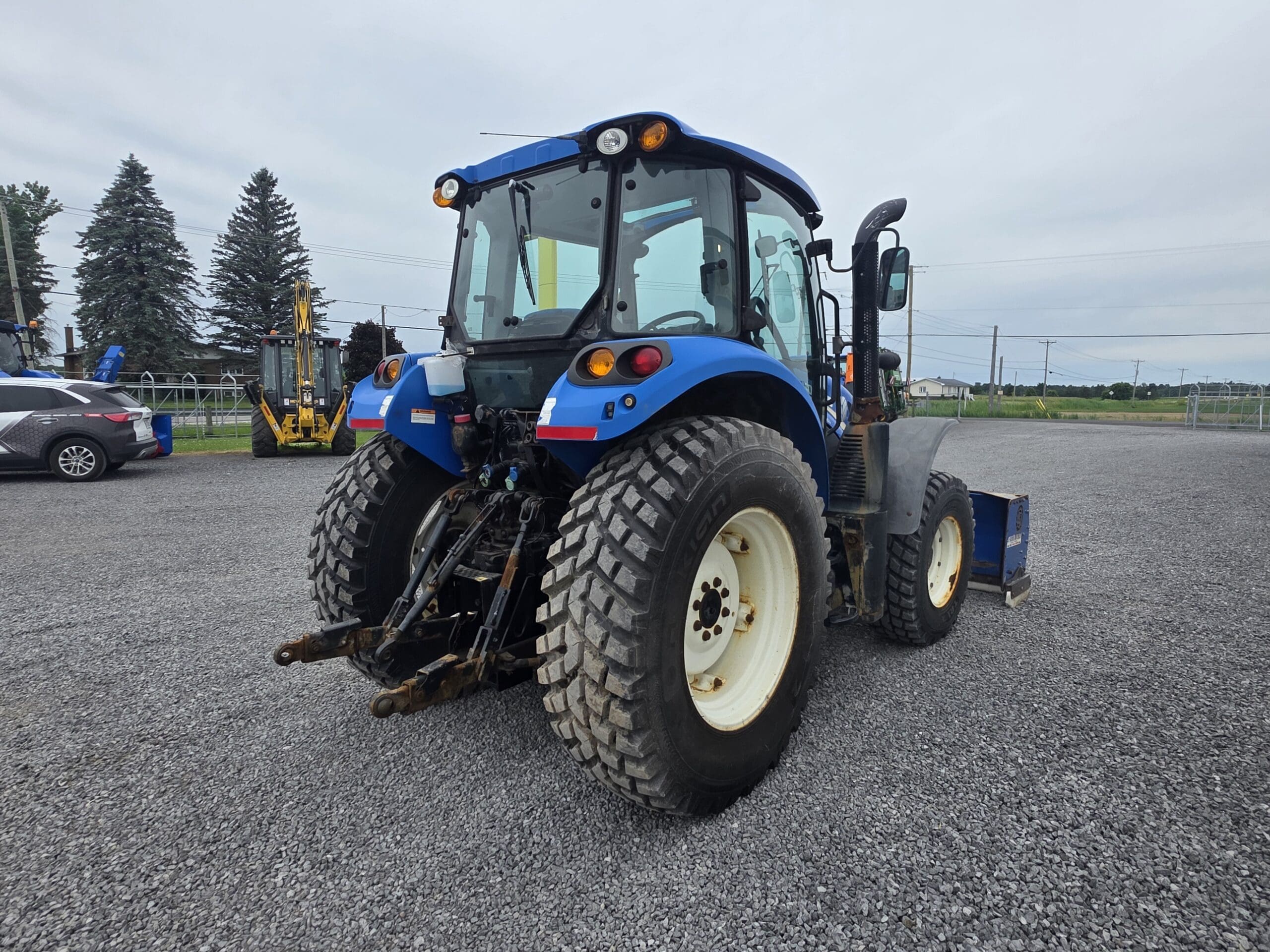 Un tracteur bleu avec de gros pneus arrière, garé sur un terrain en gravier, vu de l'arrière. À l'arrière-plan, une voiture et des engins de chantier, des arbres et un ciel nuageux.