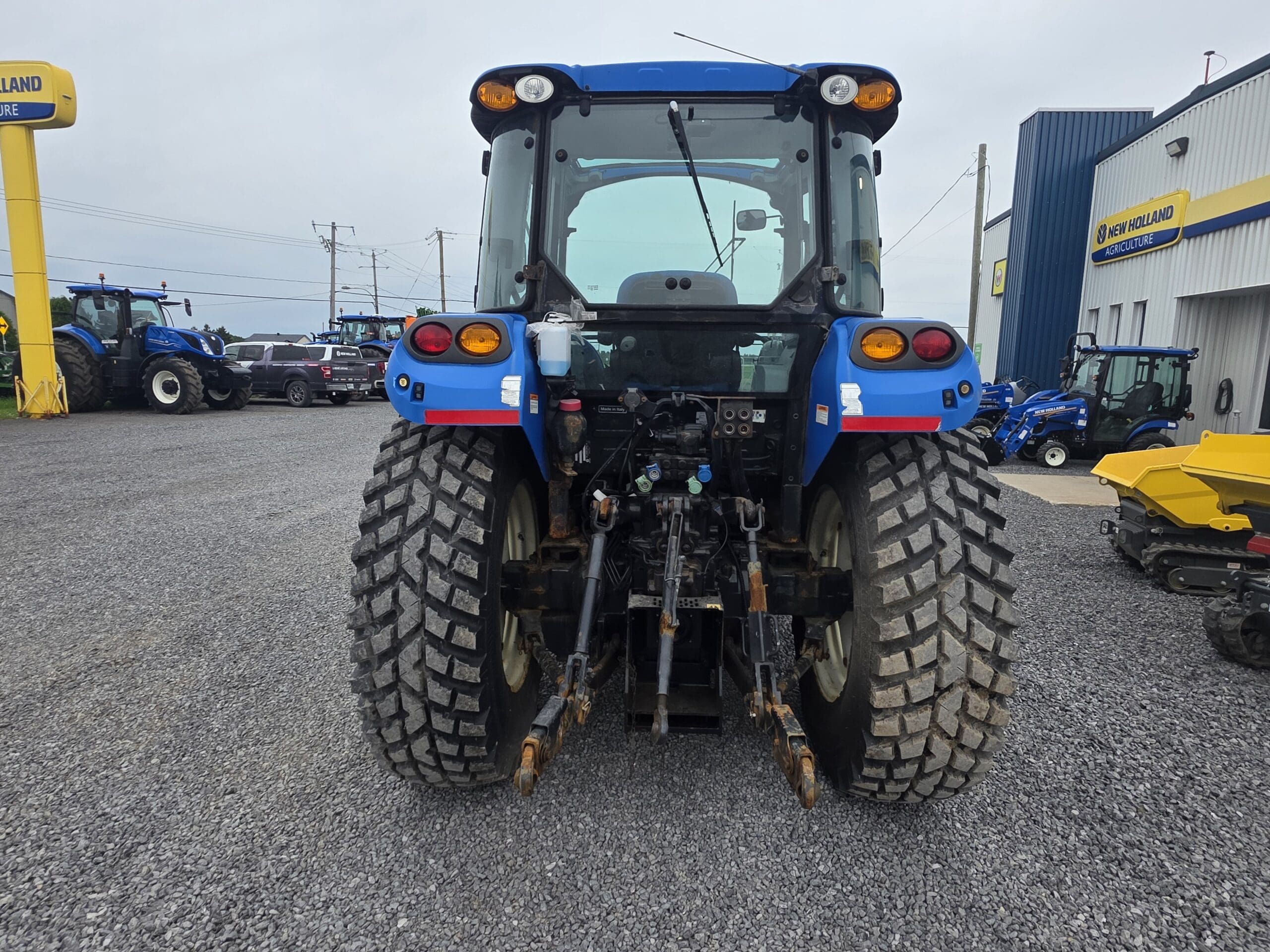 Vue arrière d'un tracteur bleu à gros pneus robustes garé sur du gravier devant le bâtiment d'un concessionnaire, avec d'autres tracteurs et équipements visibles à l'arrière-plan.
