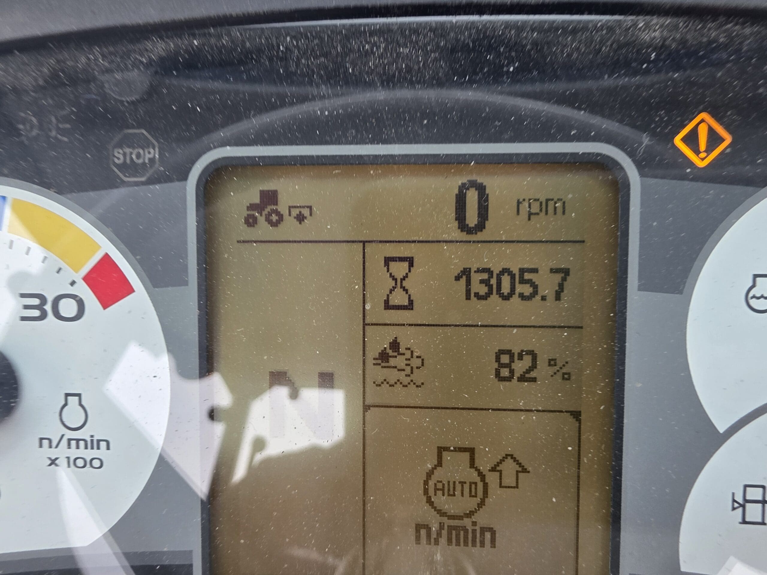 Un gros plan de l'écran du tableau de bord d'un tracteur indique 1305,7 heures, 82 % de carburant et 0 tr/min. Des icônes d'avertissement et de fonction sont visibles, avec des parties d'un tachymètre et d'autres jauges de part et d'autre.